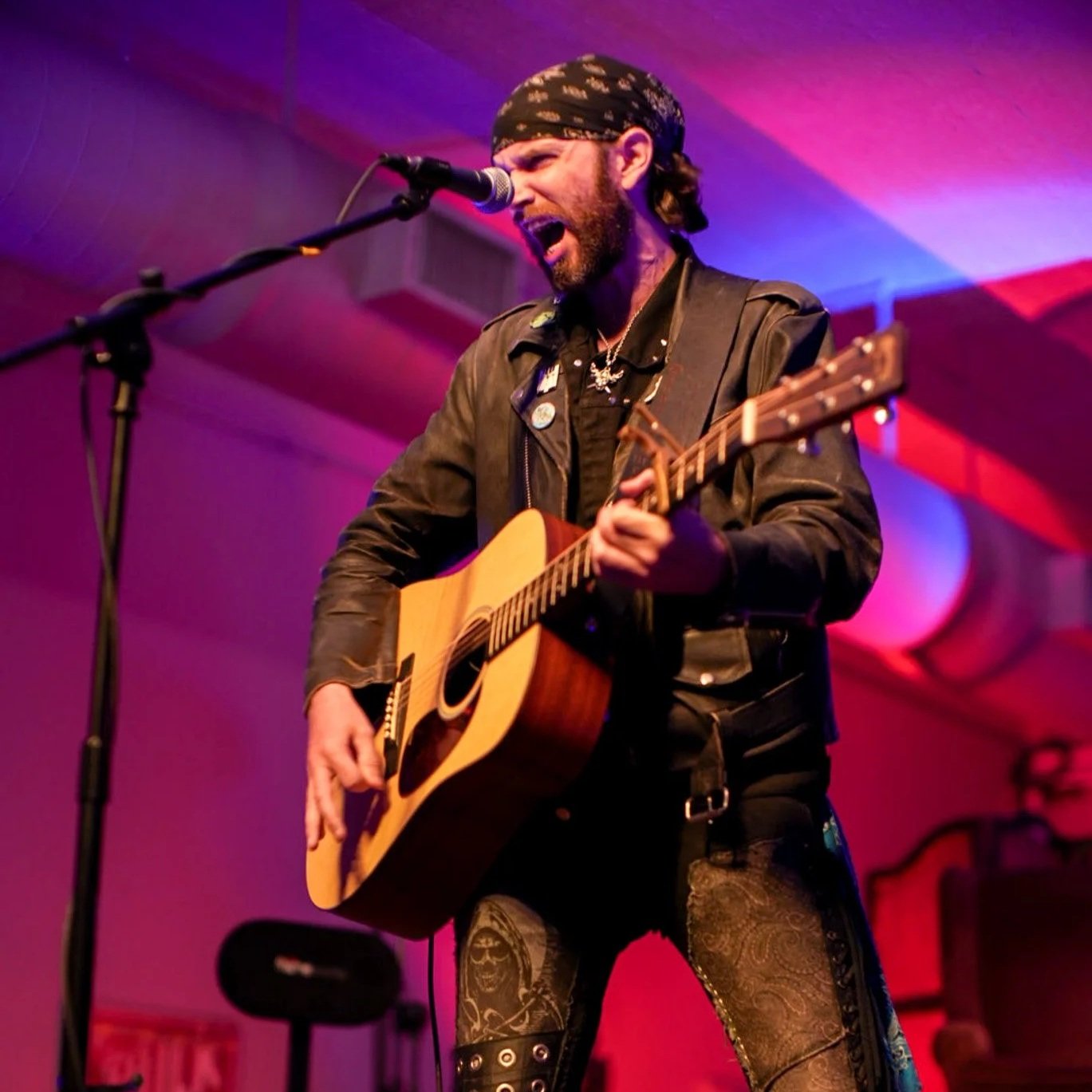 A man wearing a black bandana and leather jacket is playing an acoustic guitar and singing into a microphone on stage with red and purple lighting.