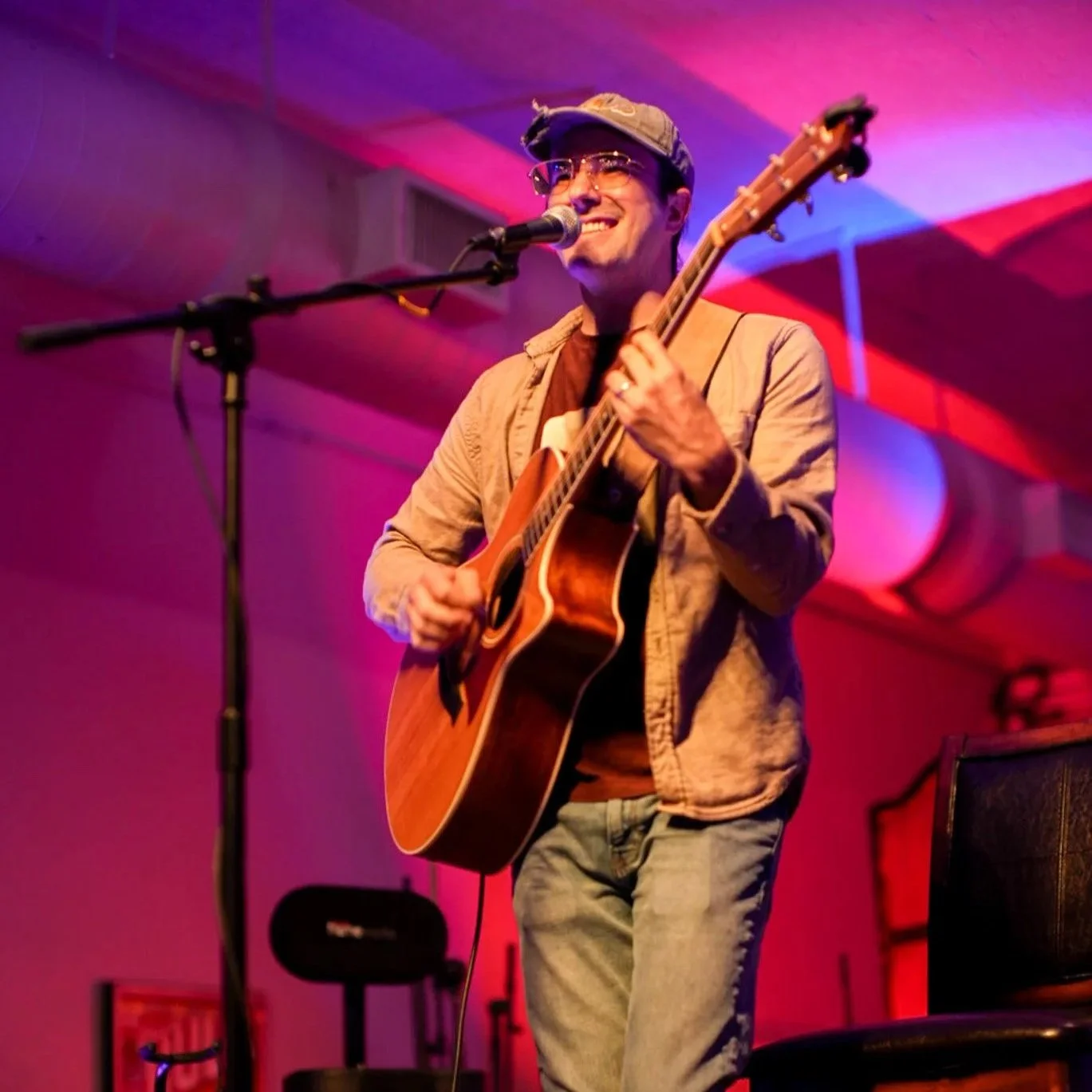 A male musician wearing glasses, a cap, and a beige jacket playing an acoustic guitar and singing into a microphone on stage with pink and purple lighting.
