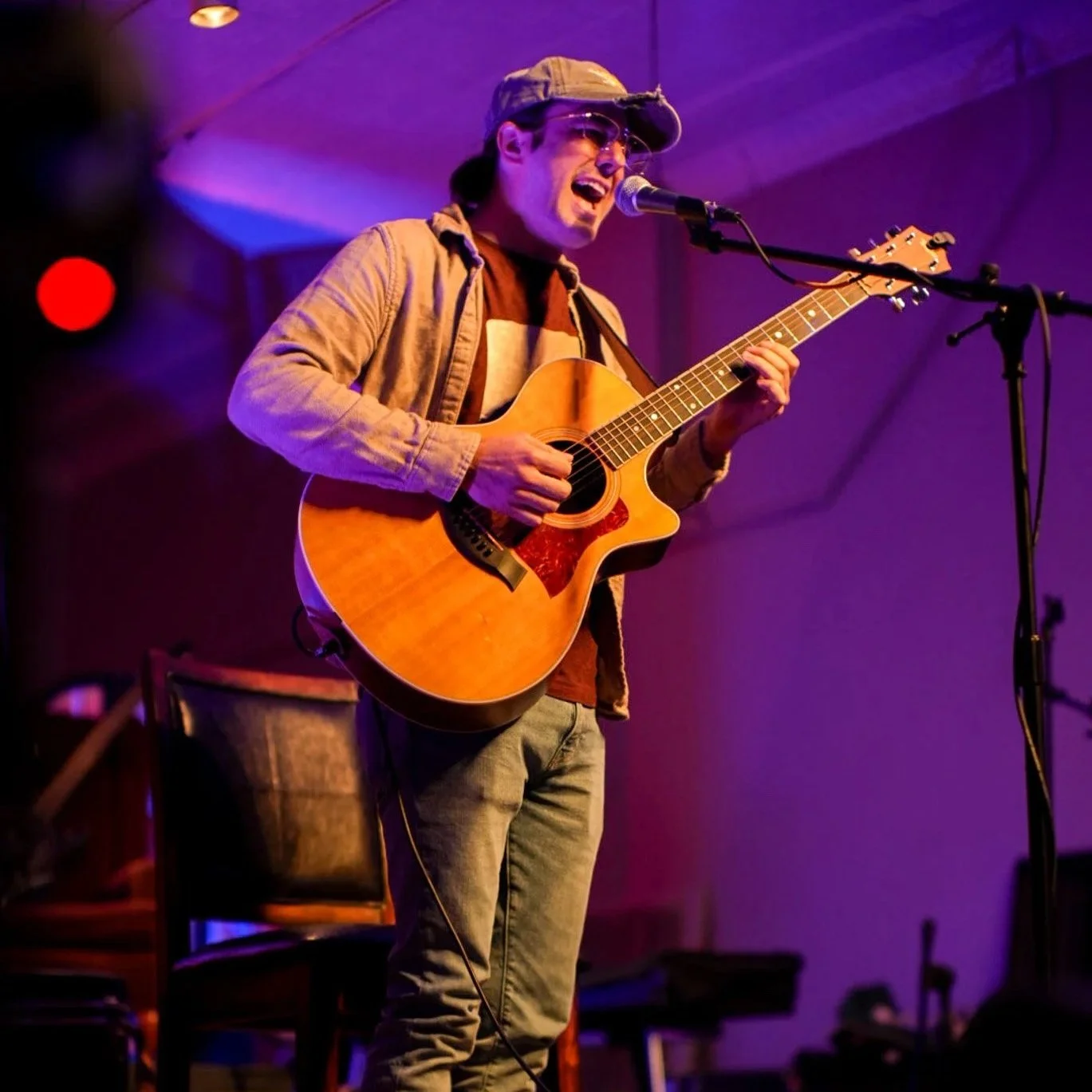 A man wearing sunglasses and a cap is playing an acoustic guitar and singing into a microphone on stage.