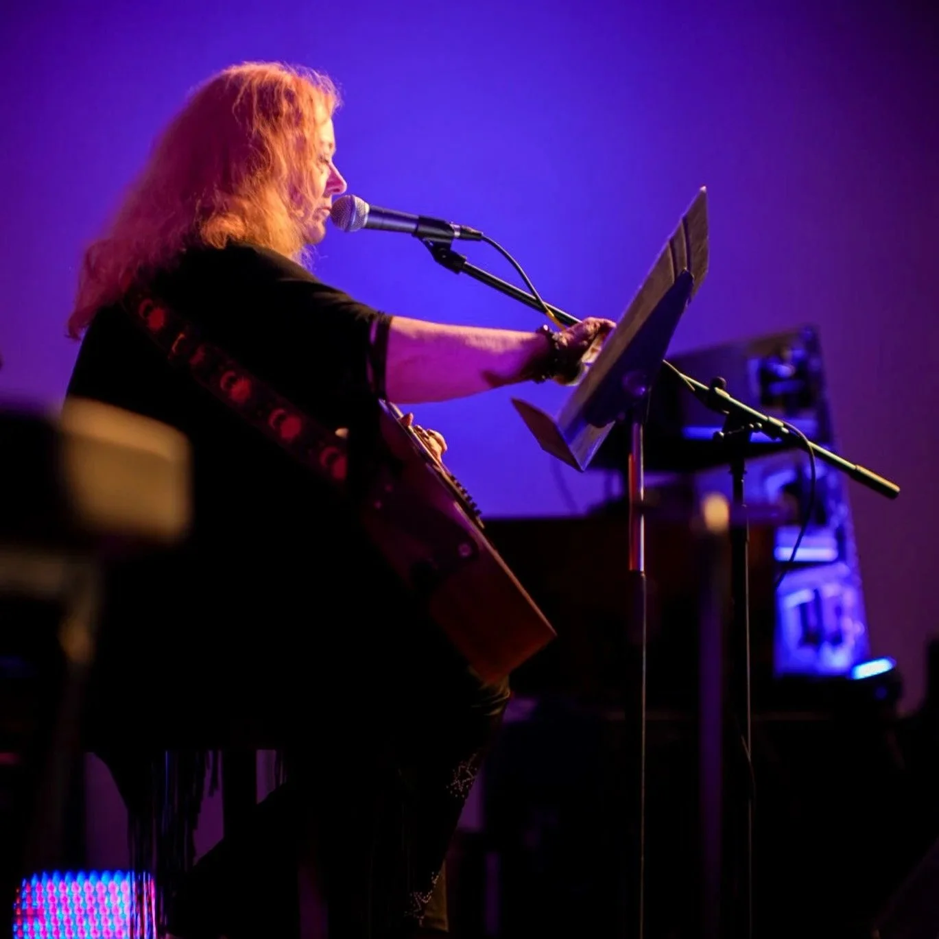A woman with red hair singing into a microphone while playing an acoustic guitar on stage. She is reading lyrics from a music stand with purple stage lighting in the background.