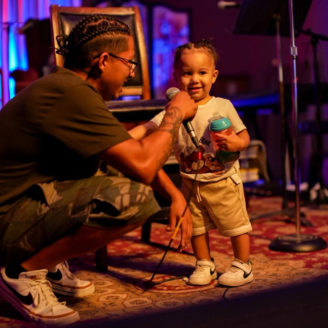 A young boy holding a water bottle and microphone, smiling at an adult with braided hair and glasses, in a dimly lit room with a musical stage in the background.