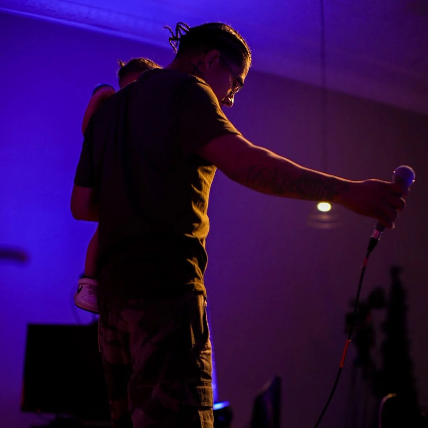 A man with glasses holding a microphone, standing on a stage with purple and blue lighting, with a child on his back.