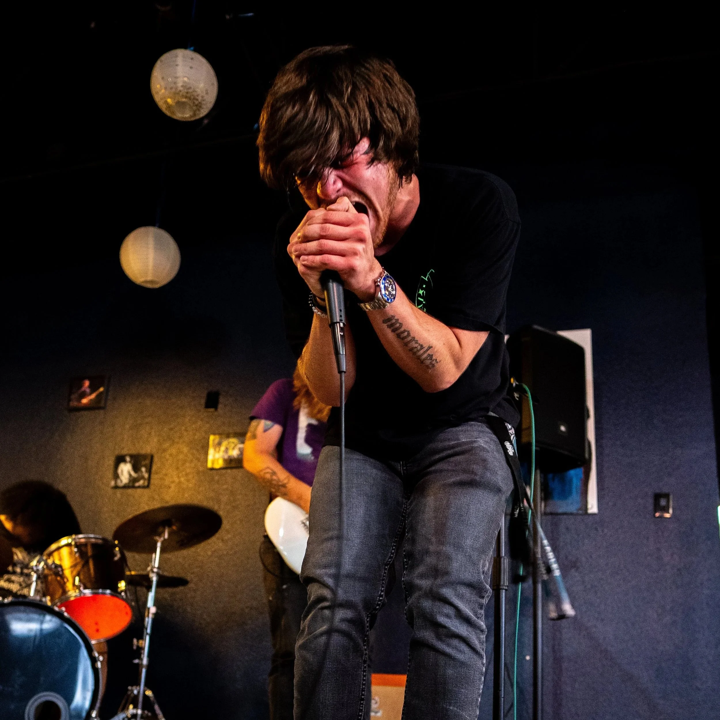 A man with brown hair singing passionately into a microphone on stage, with drum set in the background.