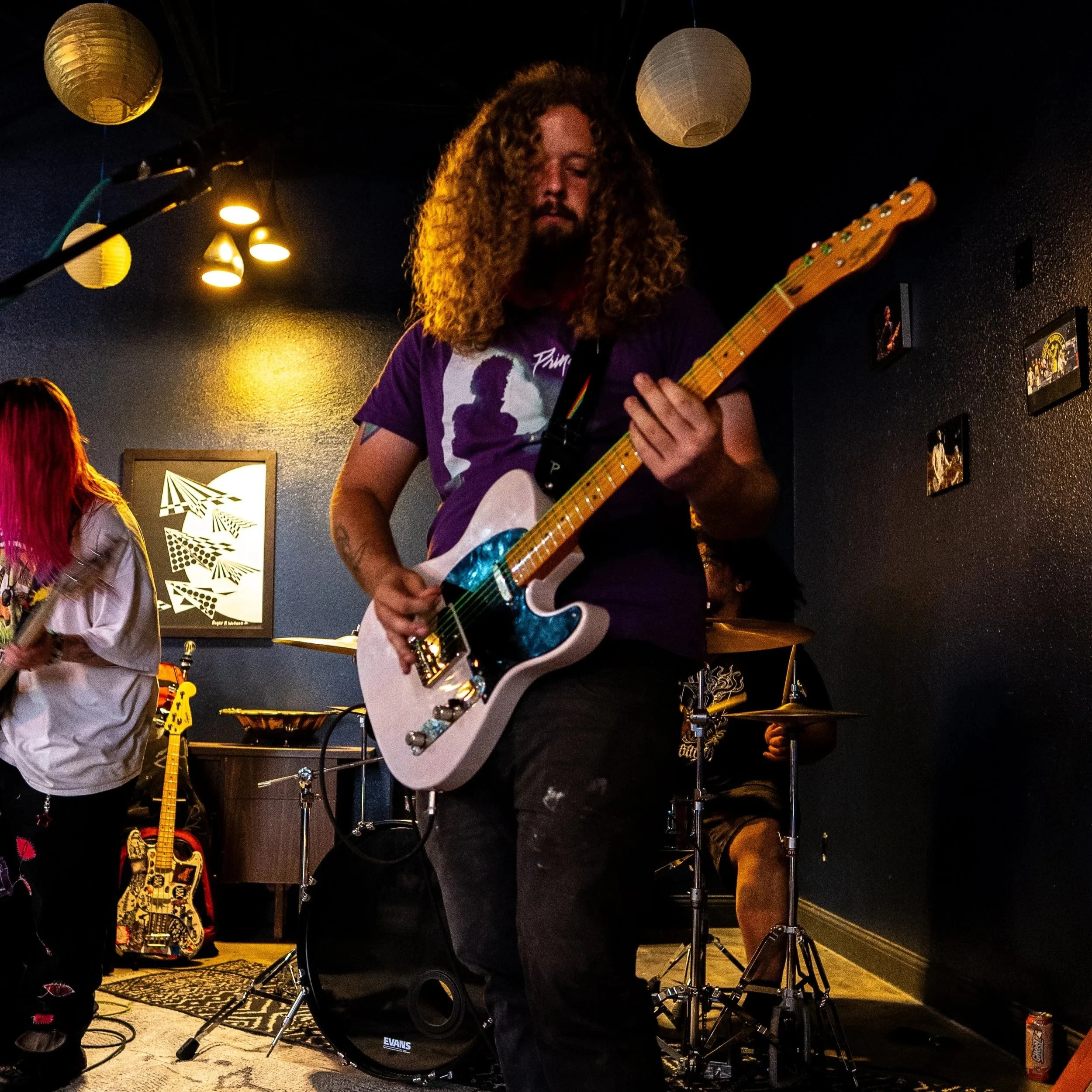 A musician with long, curly hair playing an electric guitar on stage indoors with dark walls, illuminated by warm stage lights, with a drummer's drum set and framed pictures on the wall behind him.