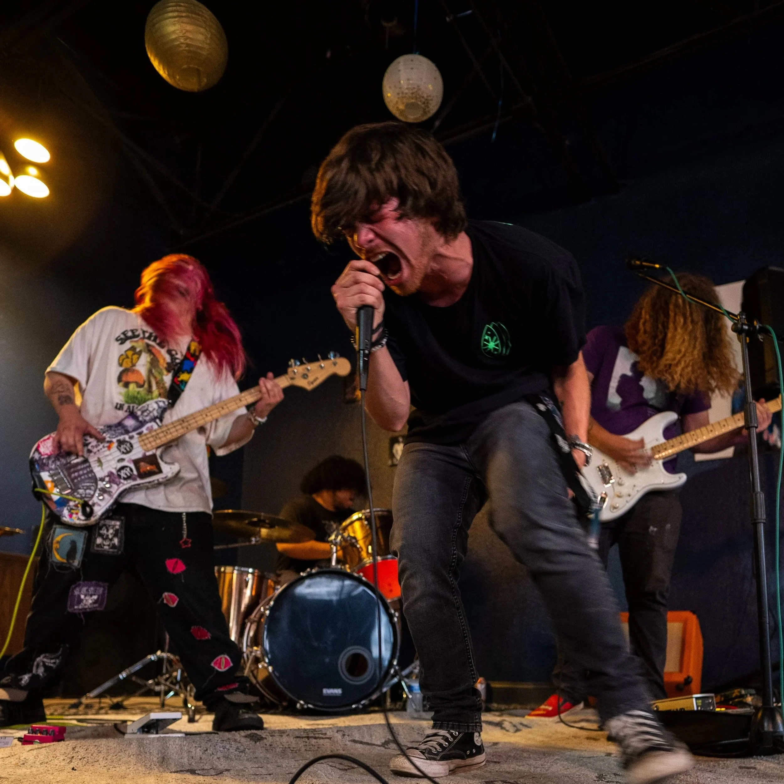 A person singing into a microphone while playing guitar on stage with band members in the background, including a woman with pink hair playing bass, indoors at a live music venue.
