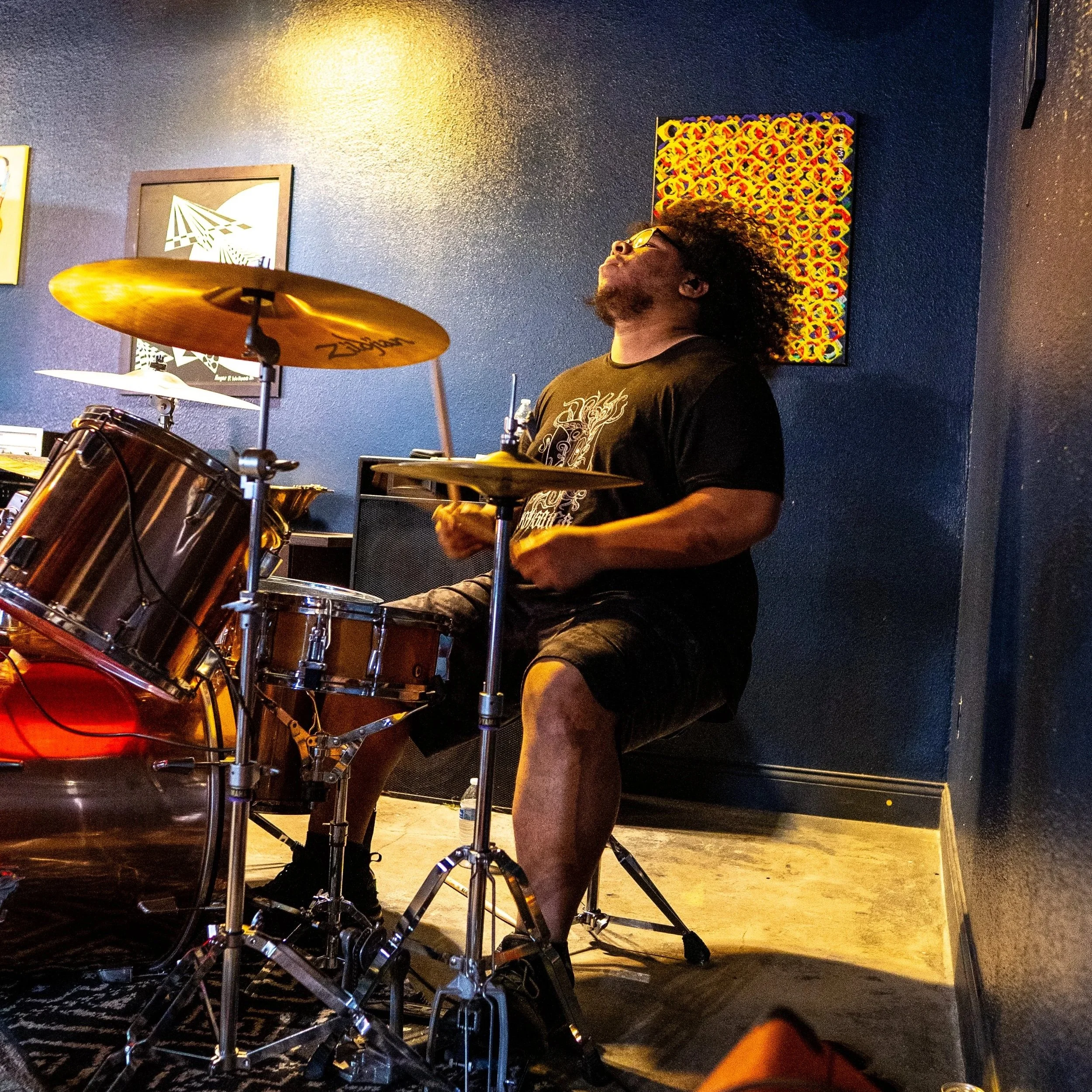 A man with curly hair and glasses playing a drum set in an indoor setting with dark blue walls and colorful artwork hanging behind him.