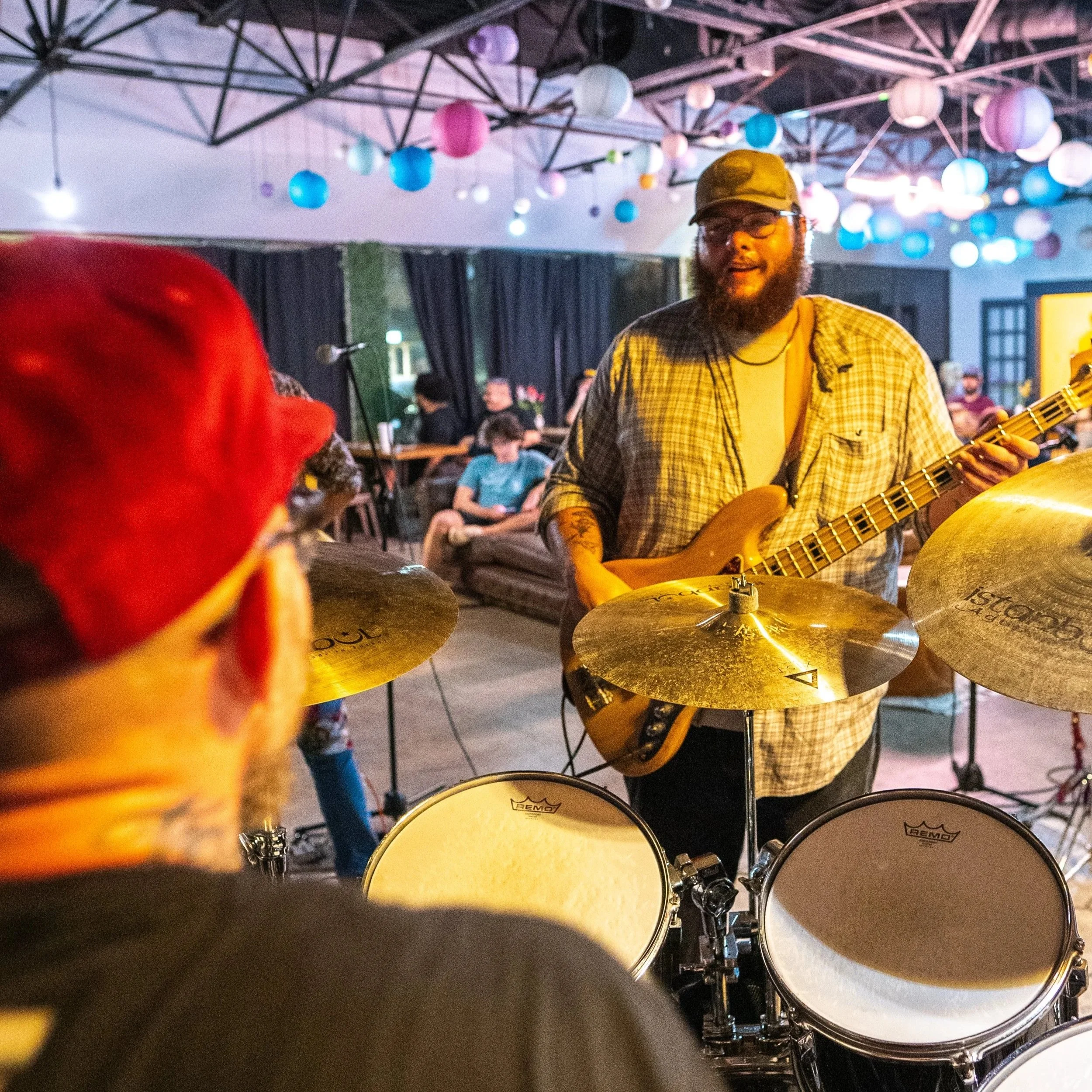 A man with a beard, glasses, and a cap, playing an electric guitar in a room decorated with colorful paper lanterns hanging from the ceiling. There are drums in the foreground and people in the background.