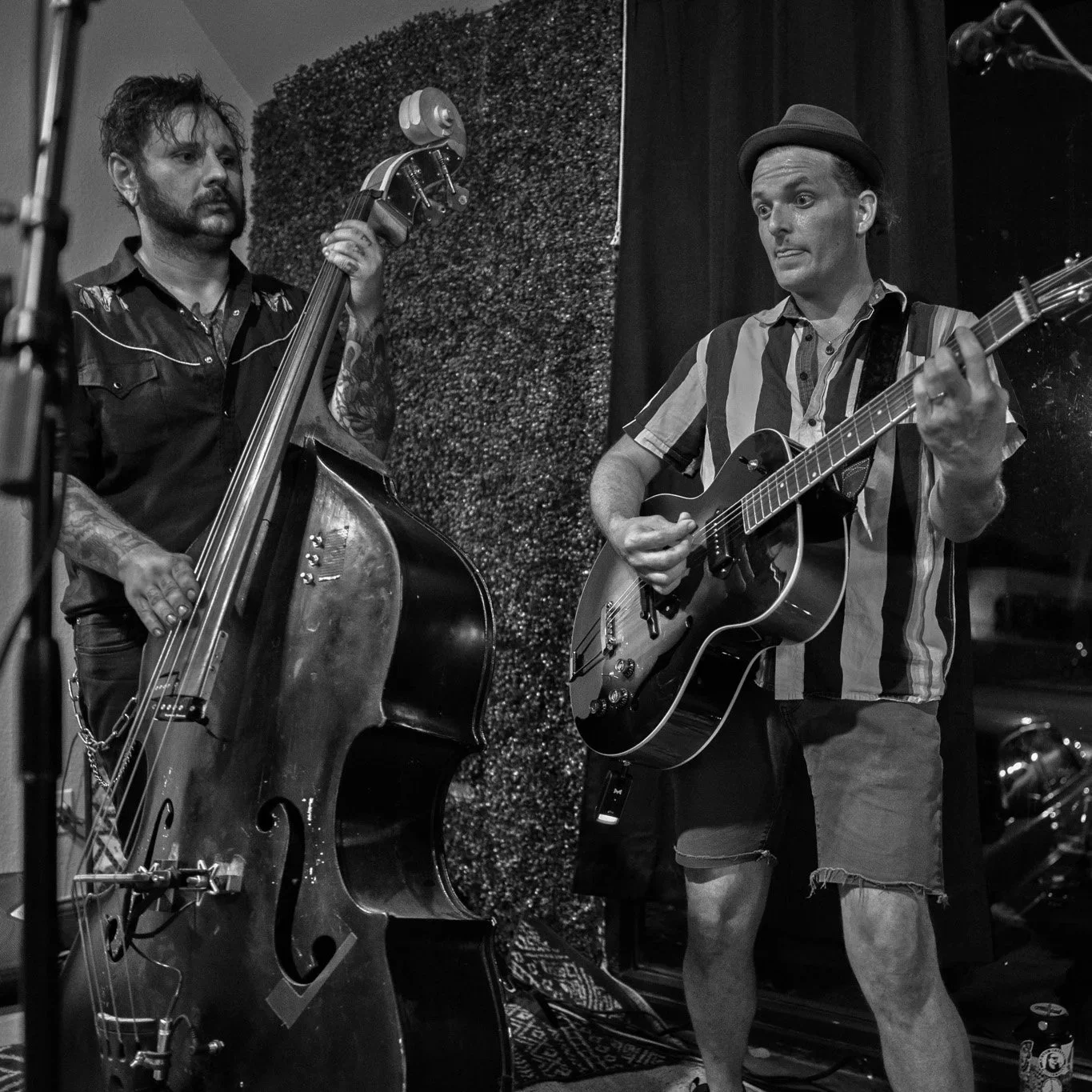 Two musicians performing with guitars, one playing an acoustic guitar and the other playing an upright bass, in a recording studio or performance space.
