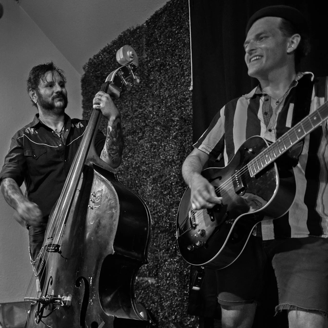 Two musicians playing instruments in a performance space, one with a double bass and the other with an electric guitar, both engaged and smiling.