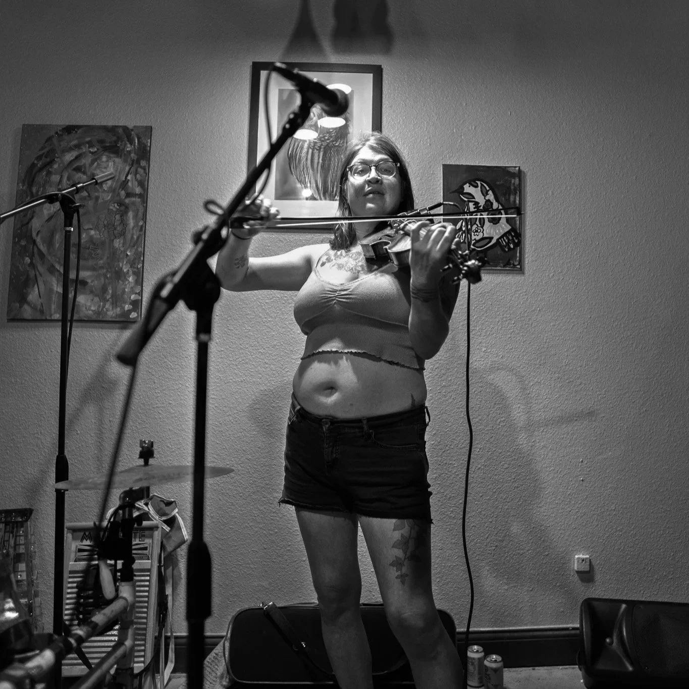 A woman with glasses and tattoos is playing a violin in a room with artwork on the walls.
