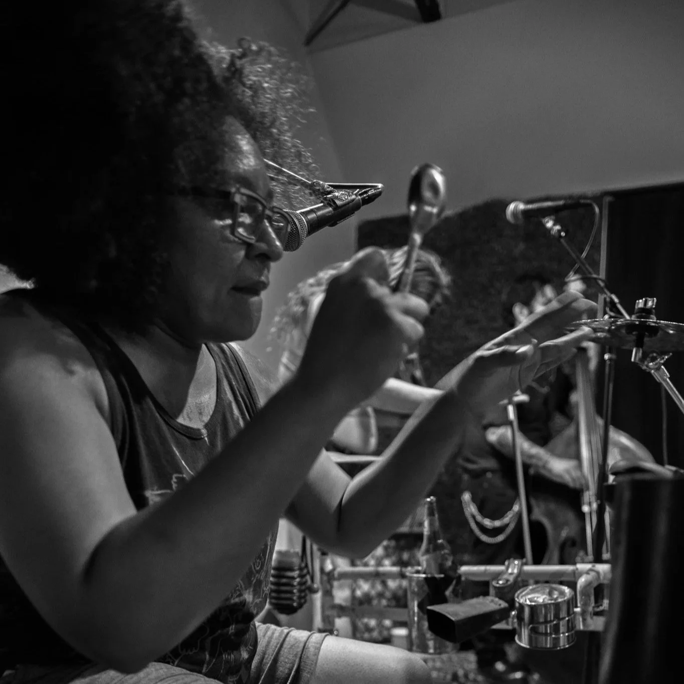 Black and white photo of a woman with curly hair and glasses, wearing a sleeveless top, playing percussion instruments on a stage with microphones and other musicians in the background.