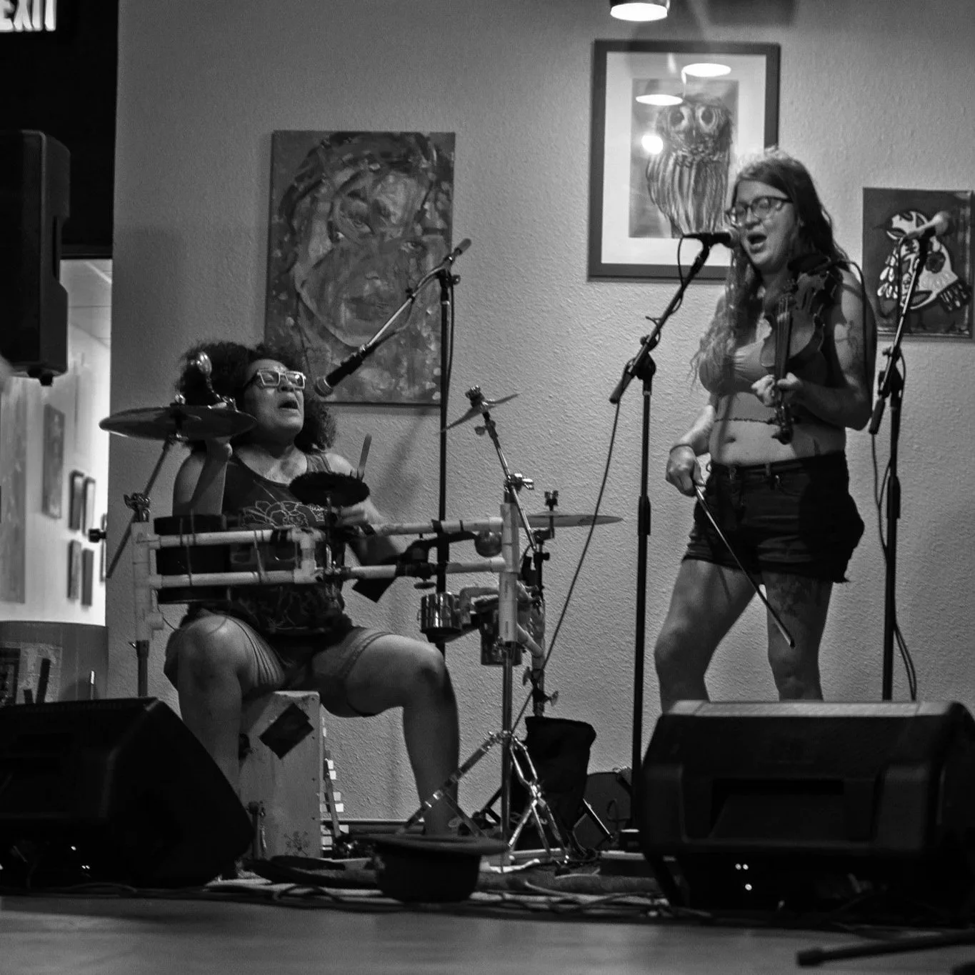 Two women performing music on a stage indoors, with one playing drums and the other singing into a microphone. Artwork hangs on the wall behind them.