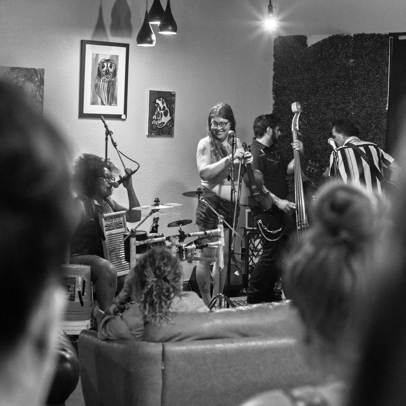 A black-and-white photo of a live music performance in a cozy indoor venue. Four musicians are on stage: a drummer with glasses, a woman with glasses smiling while holding a microphone, a man playing an upright bass, and another man with a striped sh