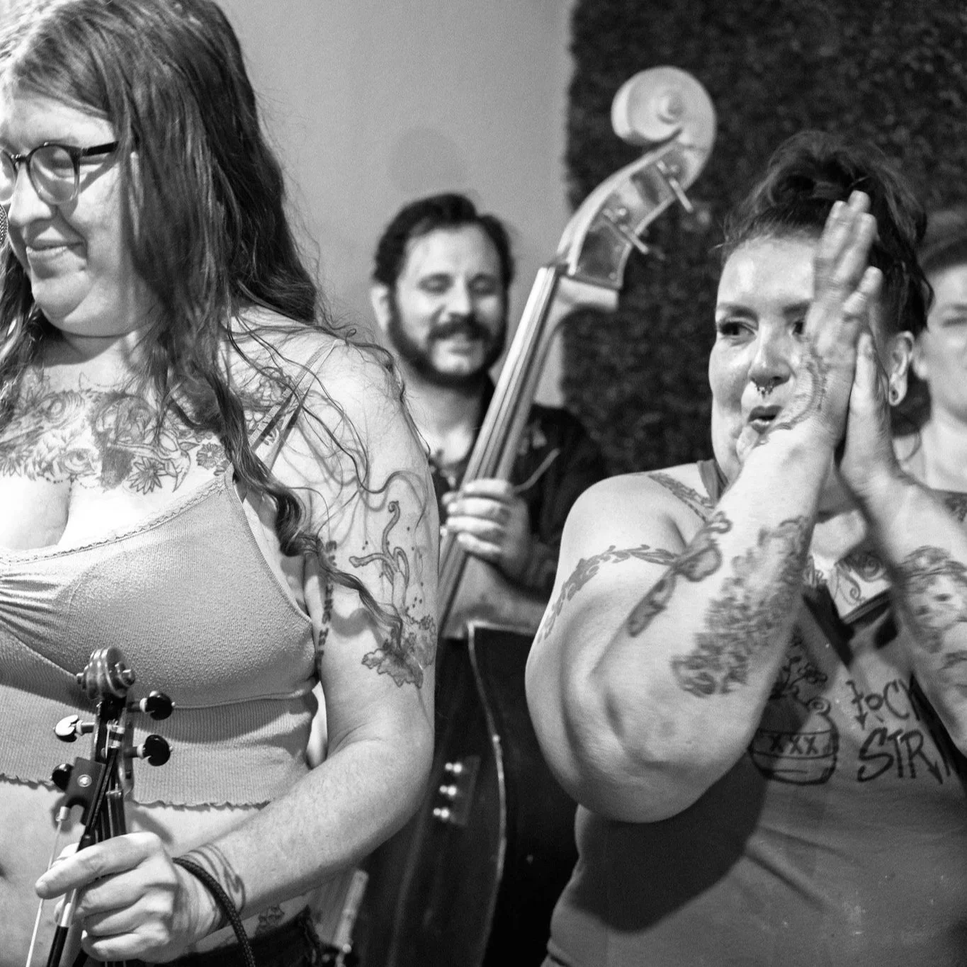 A black-and-white photo of a group of people at a musical gathering. A woman is playing a violin, smiling and looking down. Another woman is in the foreground making a peace sign near her face, with tattoos on her arms and a septum piercing. A man in