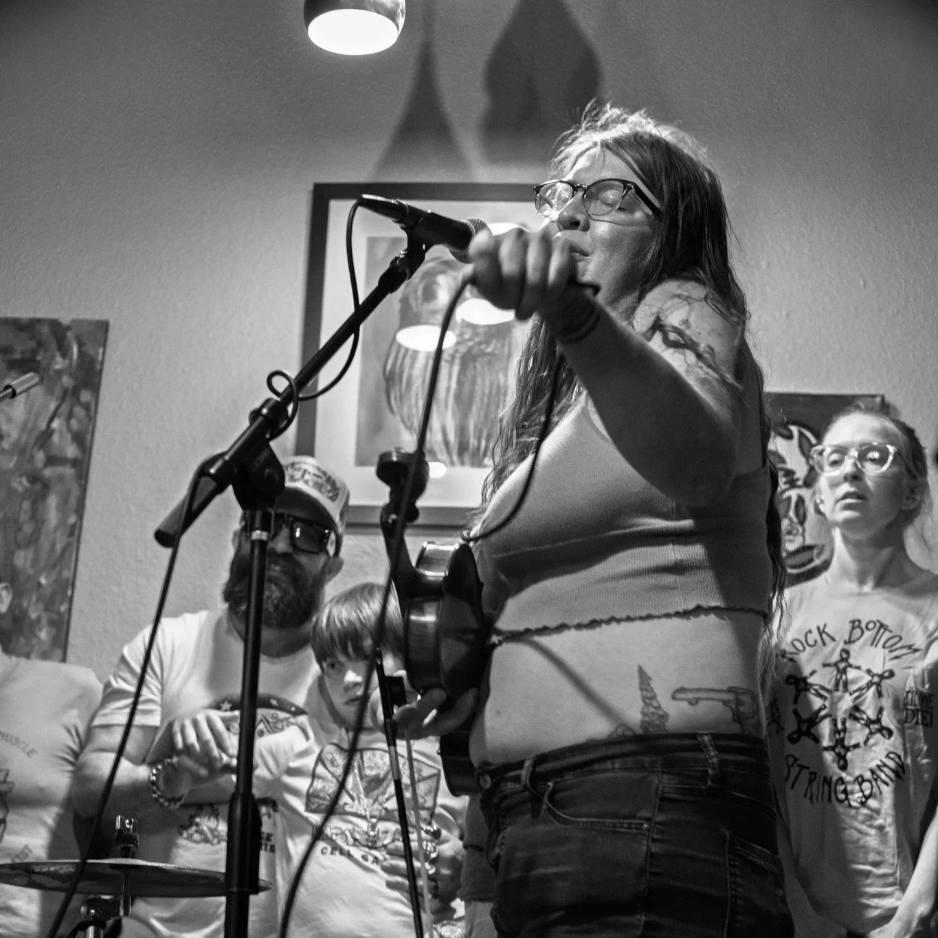 A woman singing into a microphone with glasses and long hair, surrounded by audience members in a room decorated with artwork.