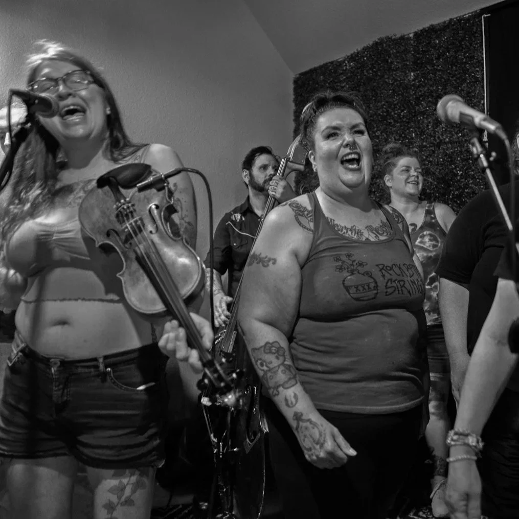 Group of women and men singing and playing instruments at a live music performance. The woman on the left is singing into a microphone and holding a violin, while the woman in the center is also singing passionately with tattoos on her arms and weari