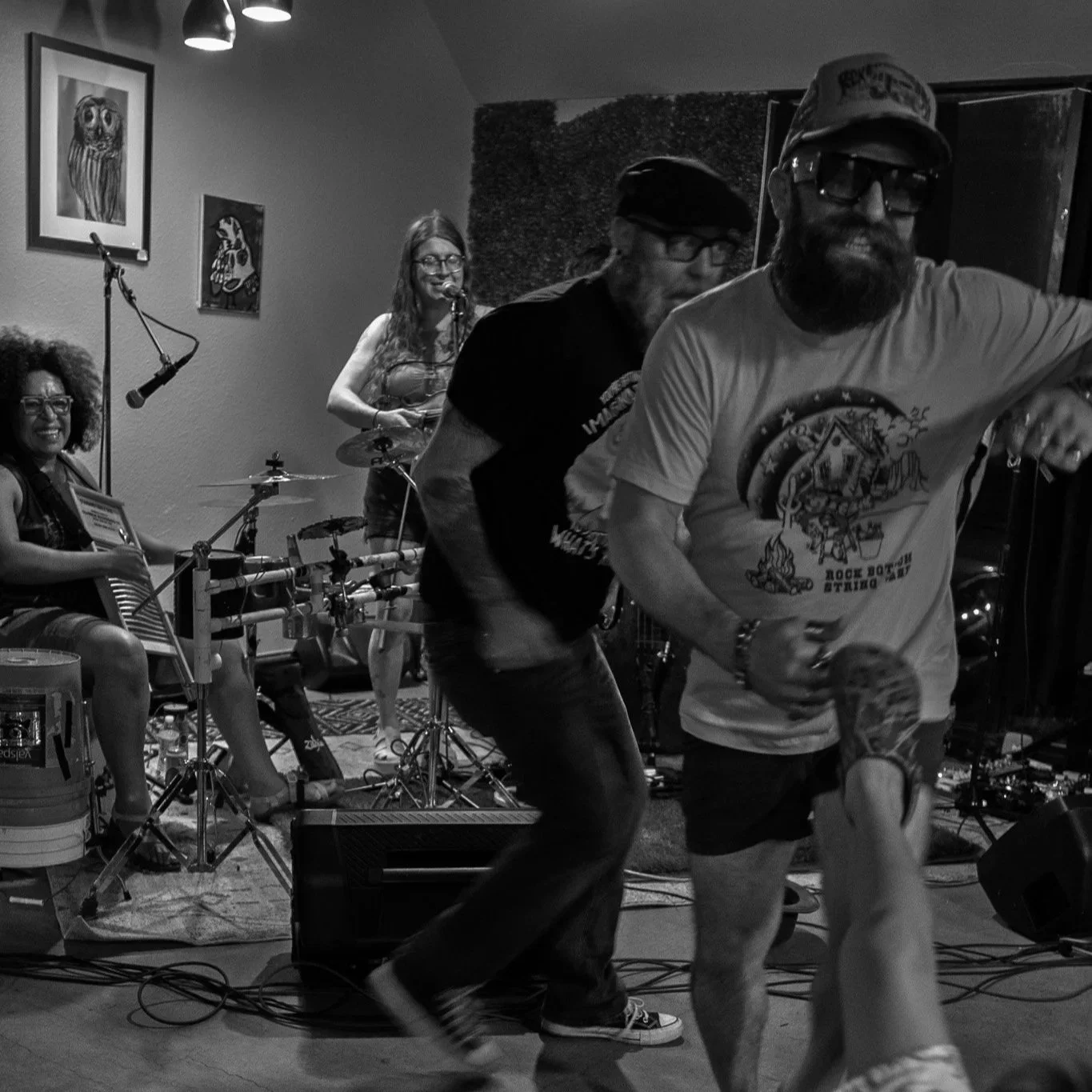 A band performing live on stage, with women playing instruments and two men dancing in front of them.