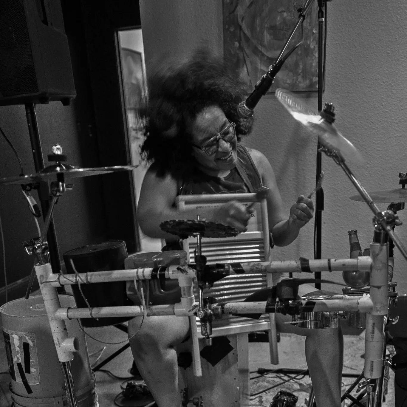 A person with curly hair and glasses, appearing excited, is surrounded by a complex structure of pipes and musical instruments like drums and cymbals, playing a percussion instrument in a room.