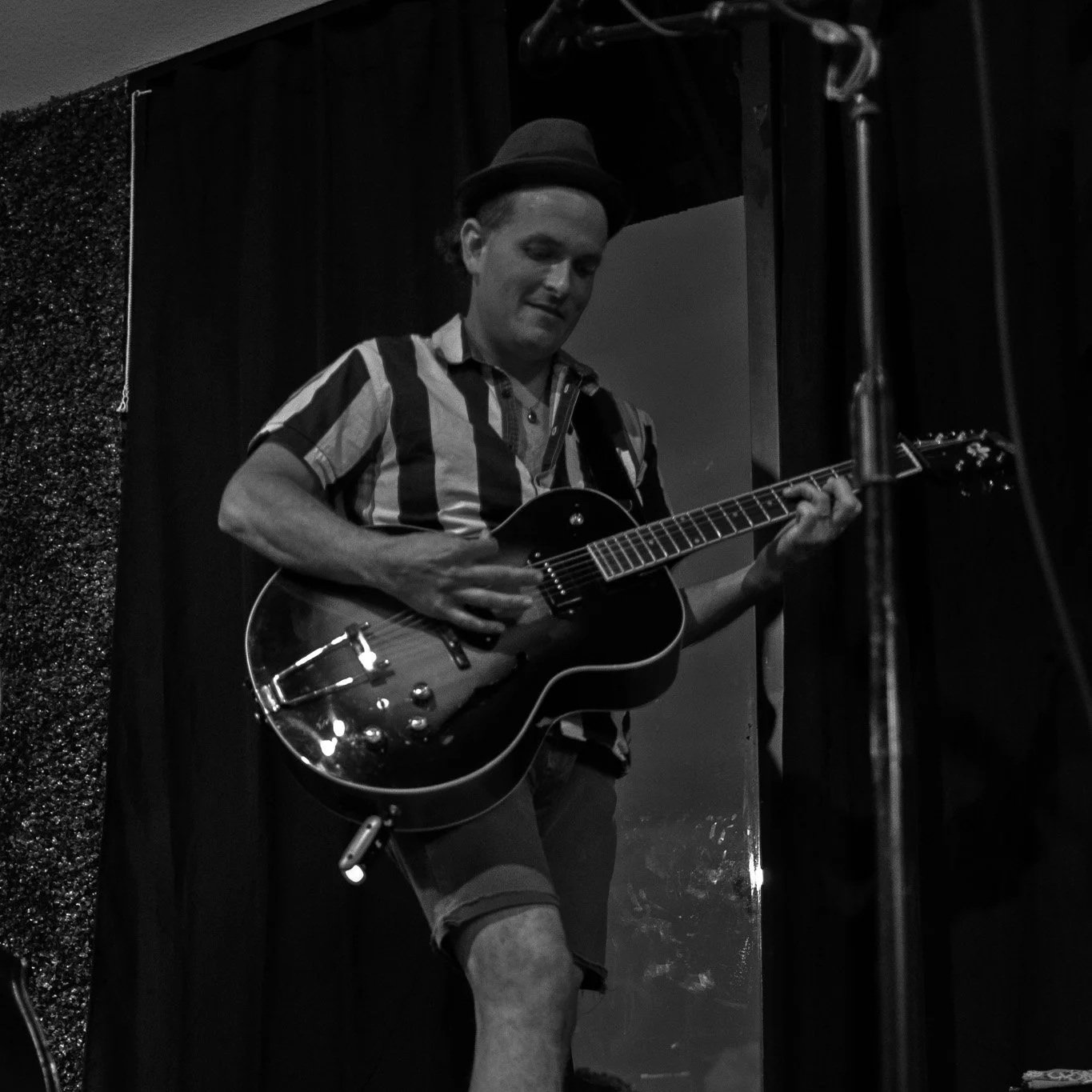 A man playing an electric guitar on stage, wearing a striped shirt, shorts, and a hat, in a black-and-white photo.