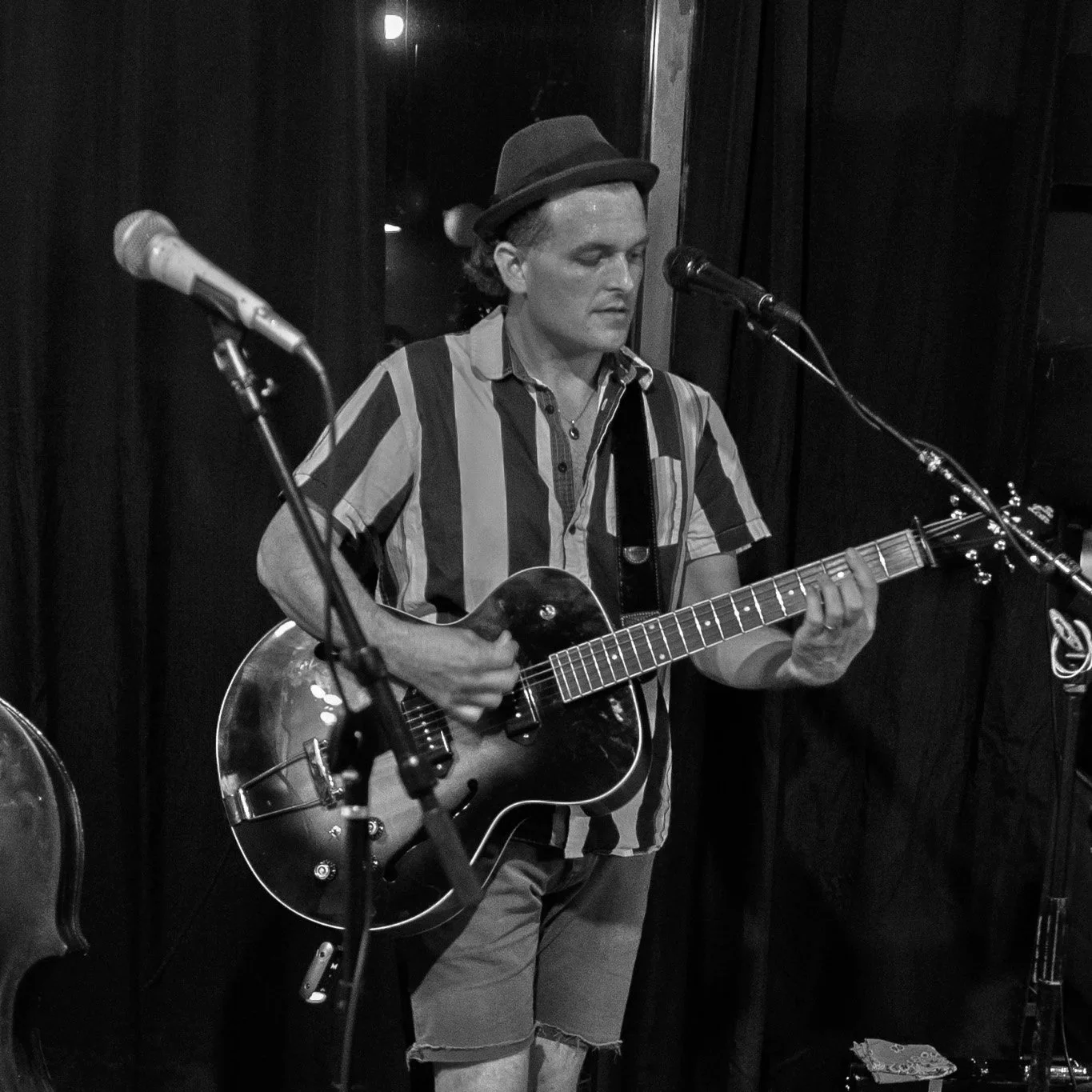A man wearing a fedora hat and striped shirt playing an acoustic guitar on stage, with microphone stands in front of him, surrounded by dark curtains.