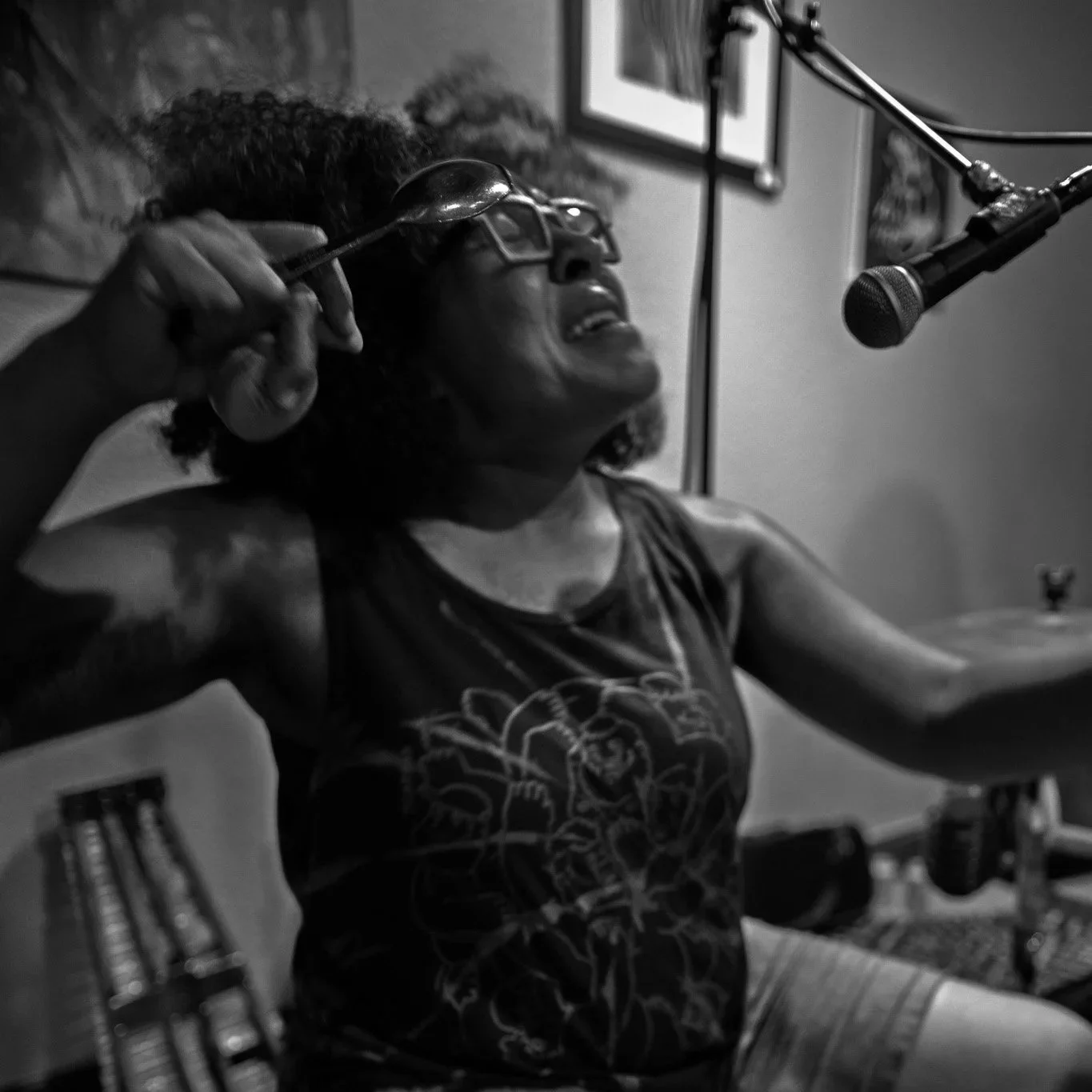 A woman singing or speaking into a microphone in a recording studio or music room. She is wearing glasses and a sleeveless shirt with a graphic design, and is holding a spoon near her head.