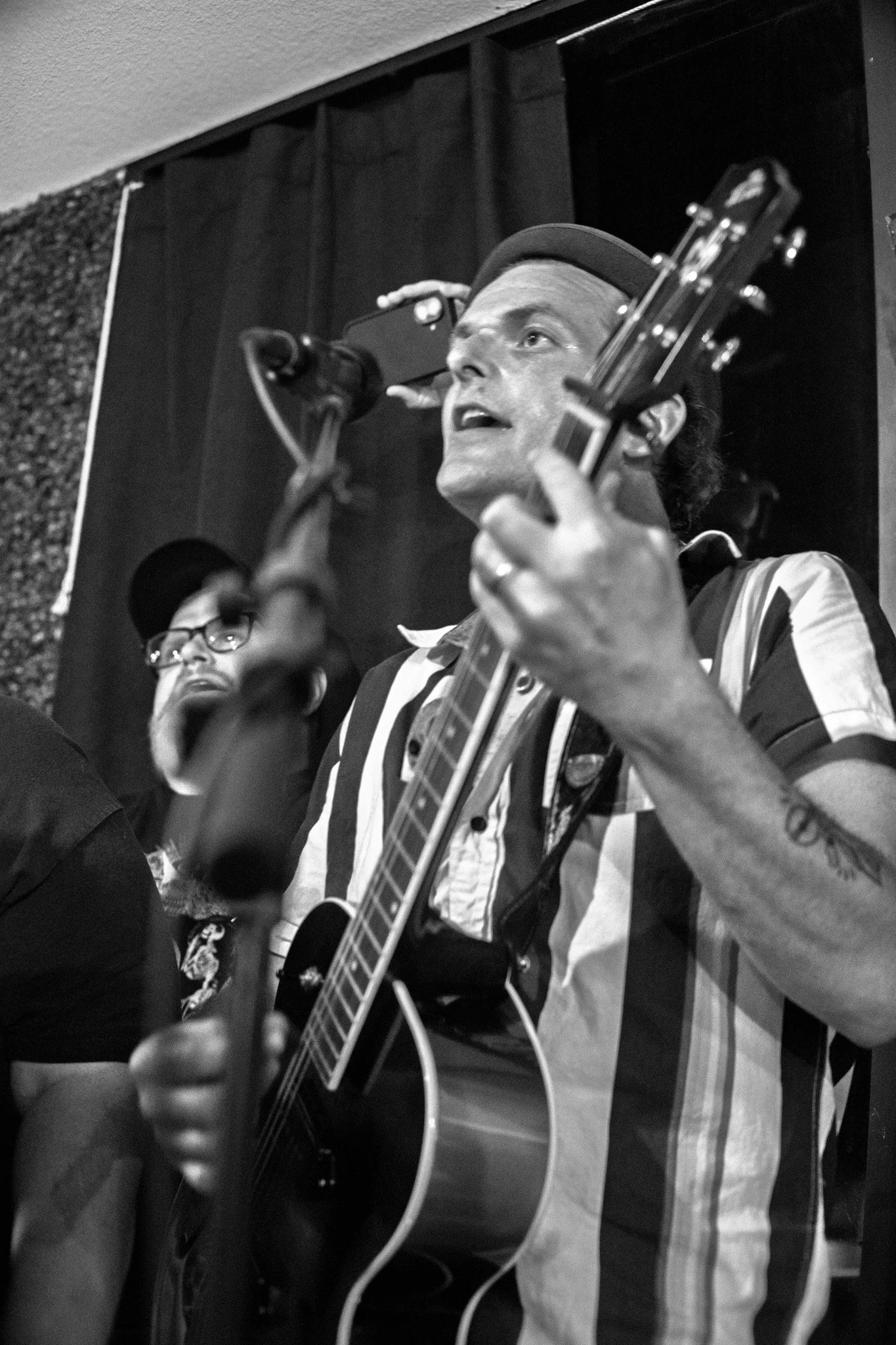 A person wearing a striped shirt and a hat, singing into a microphone while playing an acoustic guitar. Another person wearing glasses is partially visible beside him. The background includes black curtains.