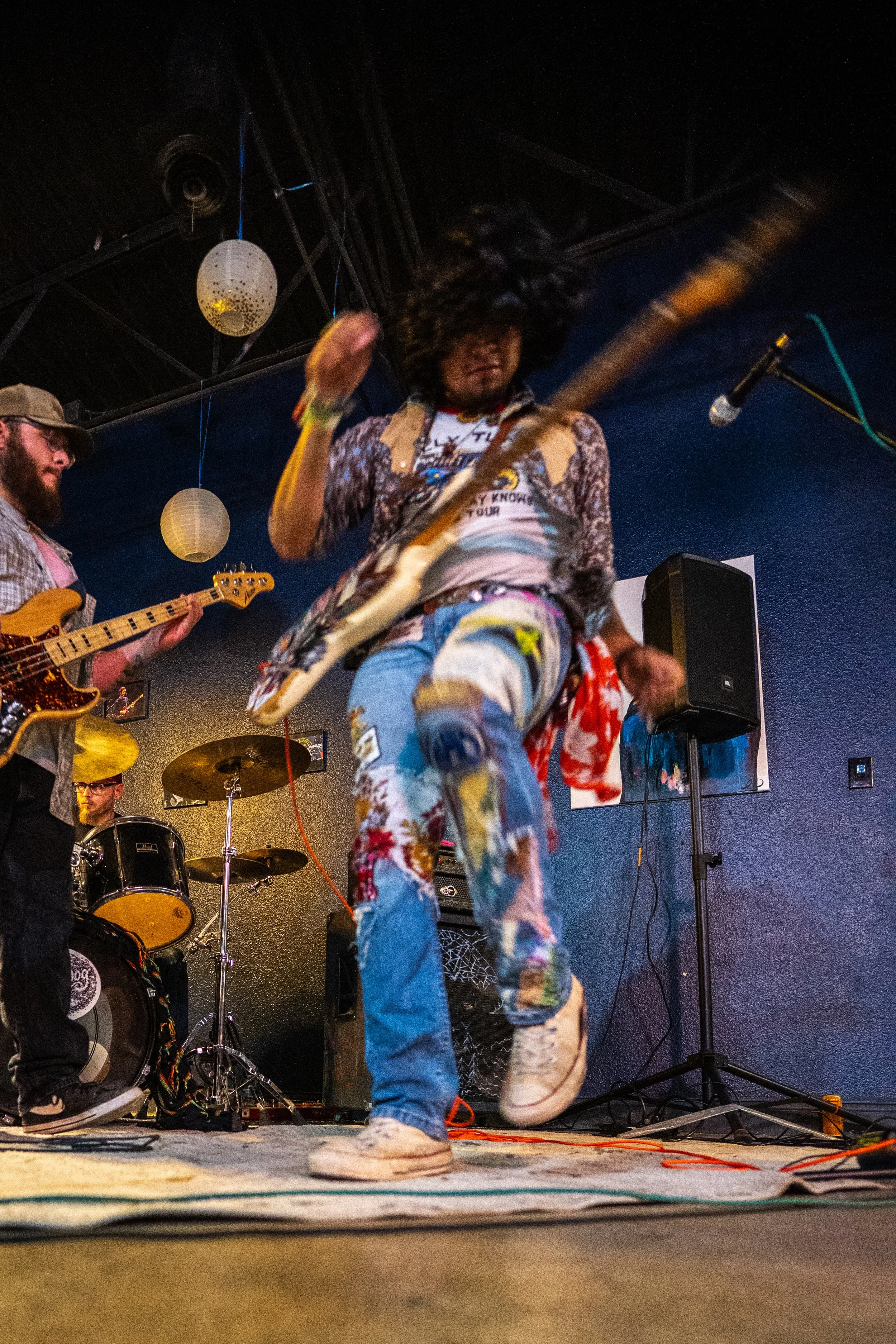 A musician with curly hair dancing and playing an electric guitar on stage, wearing colorful distressed jeans and a patchwork shirt, with a band in the background playing drums and bass.