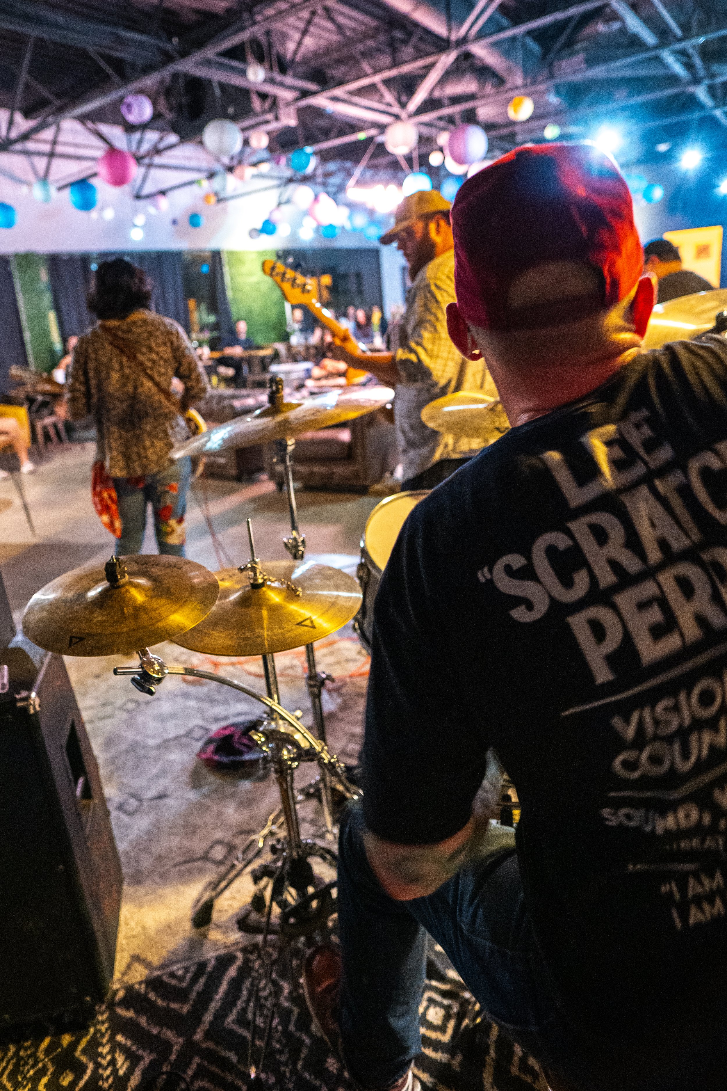 A band performing at a music venue with colorful hanging lanterns, featuring a drummer, a guitarist, and an audience in the background.