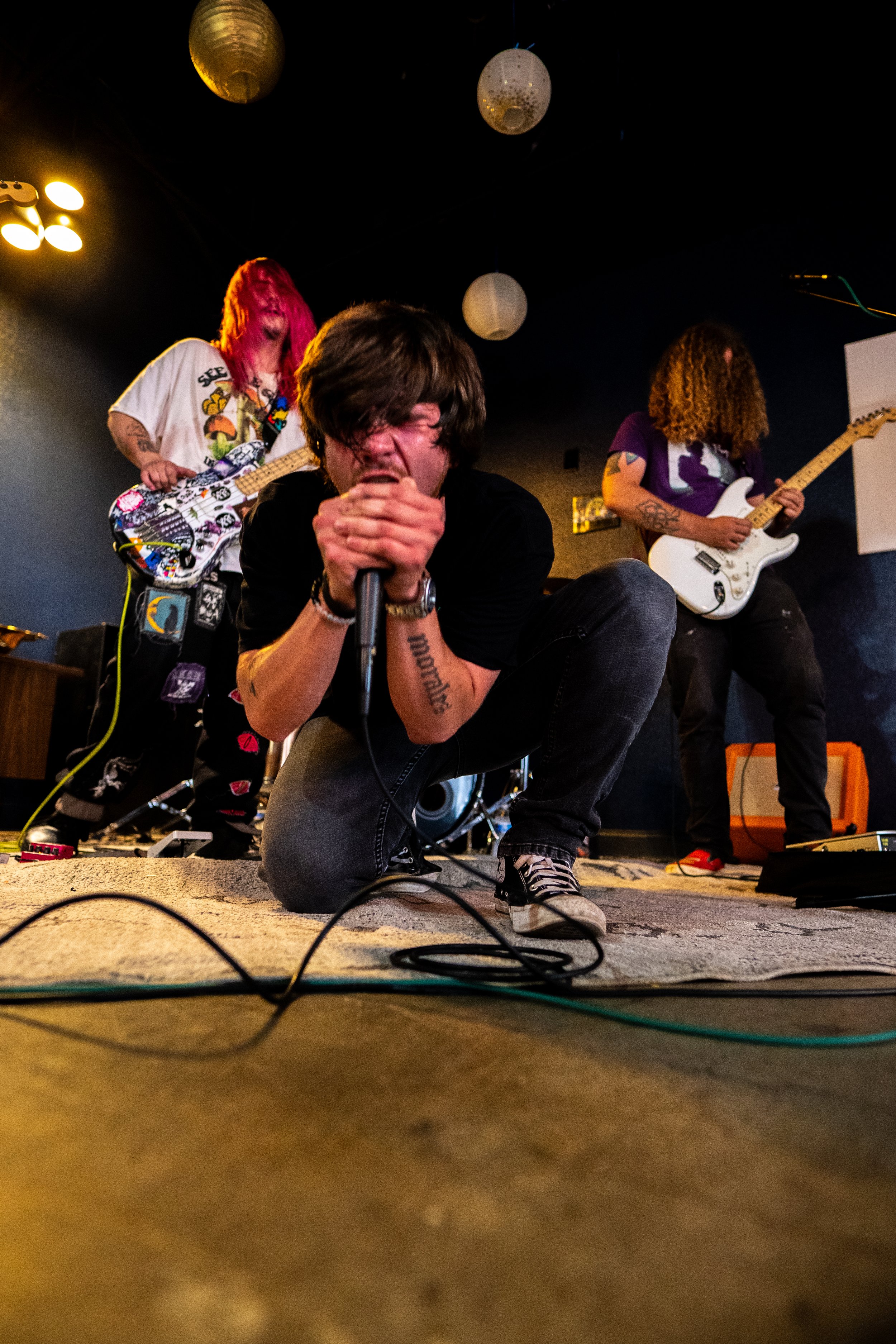 A band performing live on stage, with a singer crouching in front, holding a microphone, and two guitarists playing in the background. The stage is decorated with paper lanterns hanging from the ceiling.