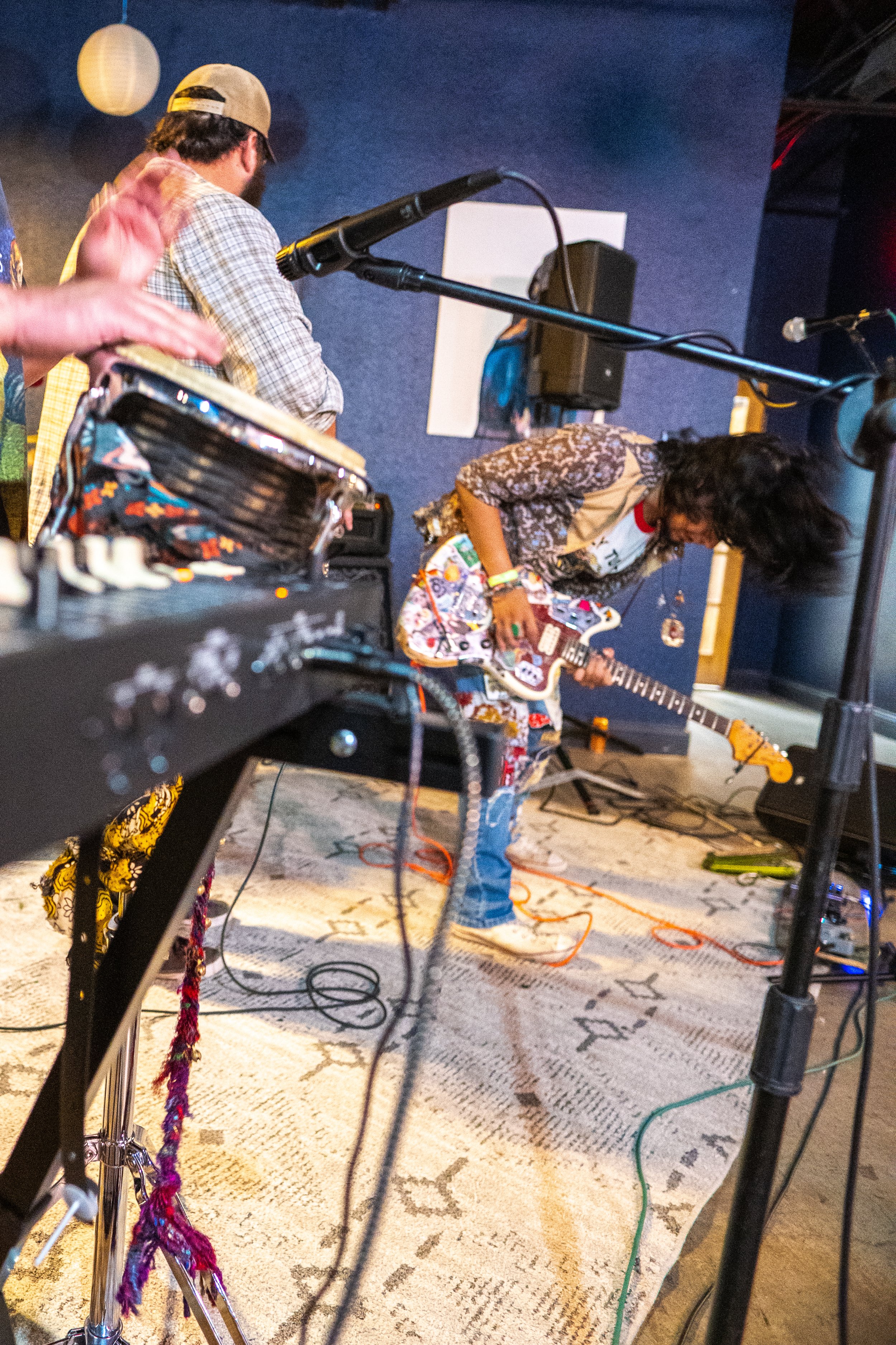 Musicians performing on stage with one person playing an electric guitar and others near a microphone and equipment, in an indoor setting.