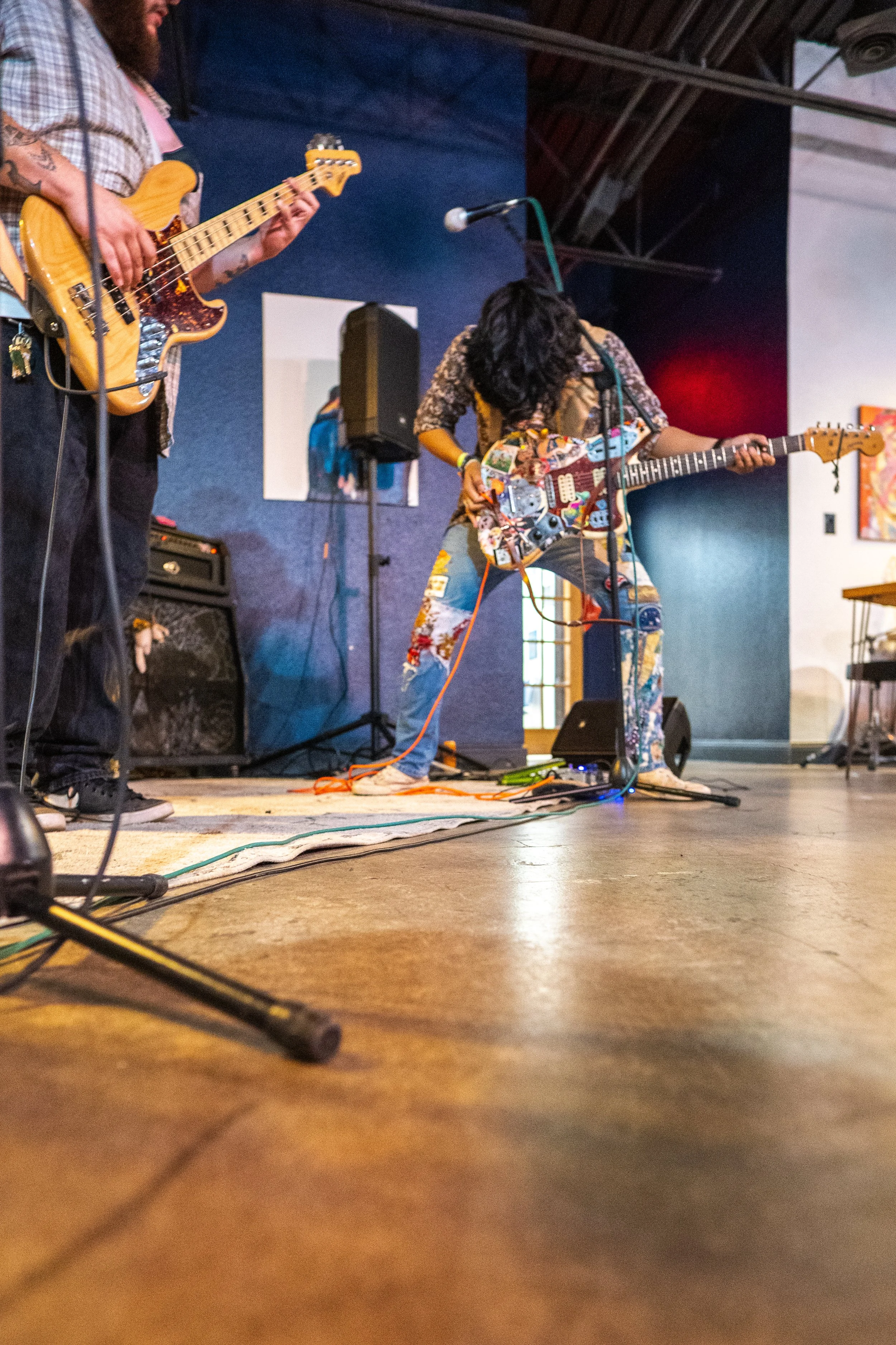Two musicians performing on stage with guitars, a person with curly hair playing a colorful electric guitar and another person playing a bass guitar, microphones, speakers, and artwork on the wall, in an indoor setting.