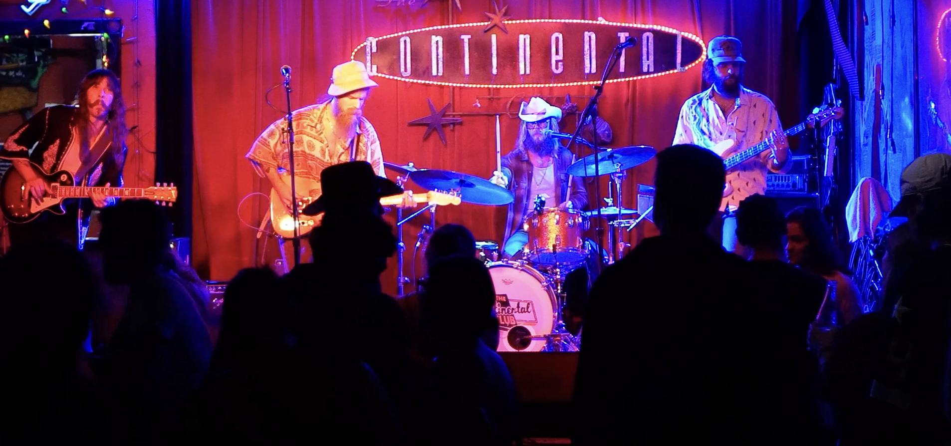 A live music performance on stage with four musicians: a guitarist on the left, a guitarist in the middle, a drummer in the center, and a bassist on the right, with a red curtain background and a sign that reads 'Continental' above the stage. Audience silhouettes are visible in the foreground.