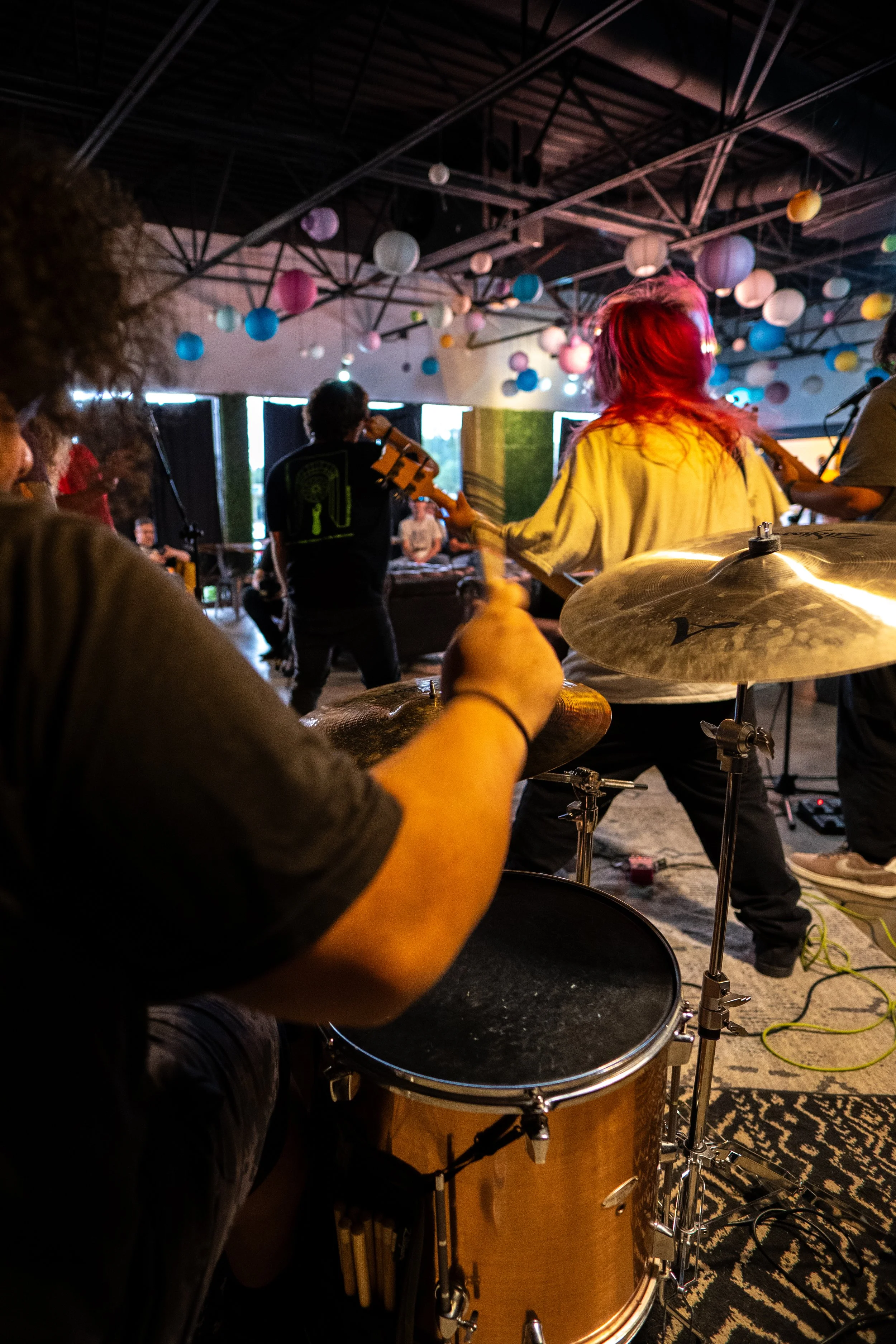 View from a drum set showing a band of musicians performing indoors, with colorful paper lanterns hanging from the ceiling.