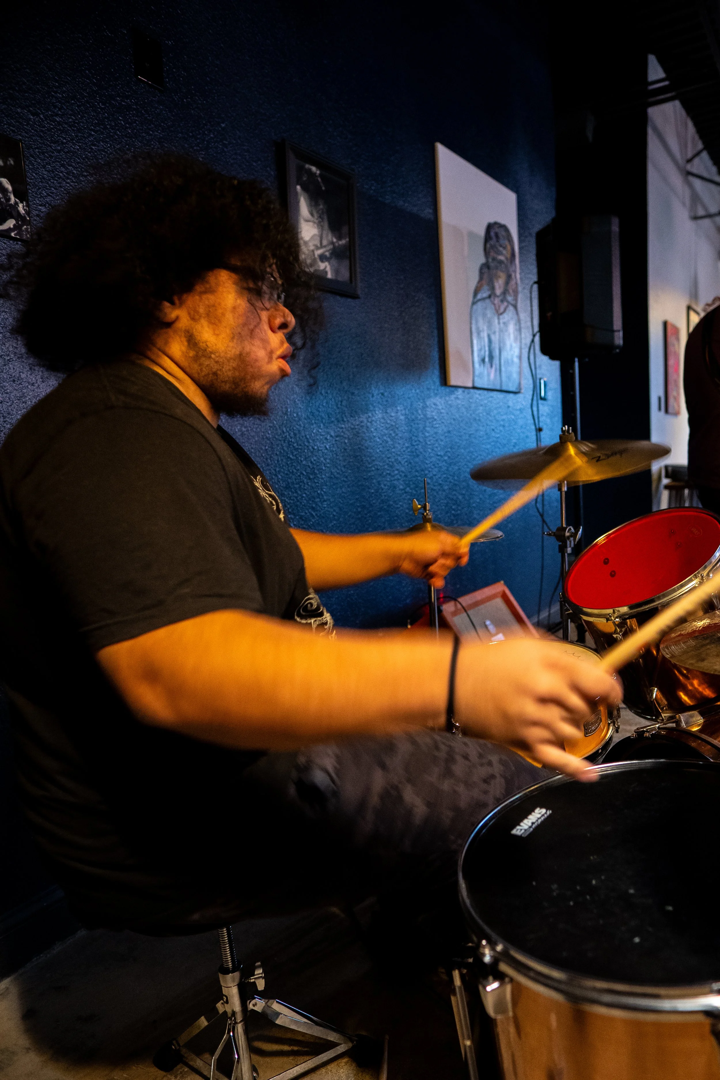 A person with curly hair playing a drumset in a dimly lit room with artworks on the dark blue wall behind him.