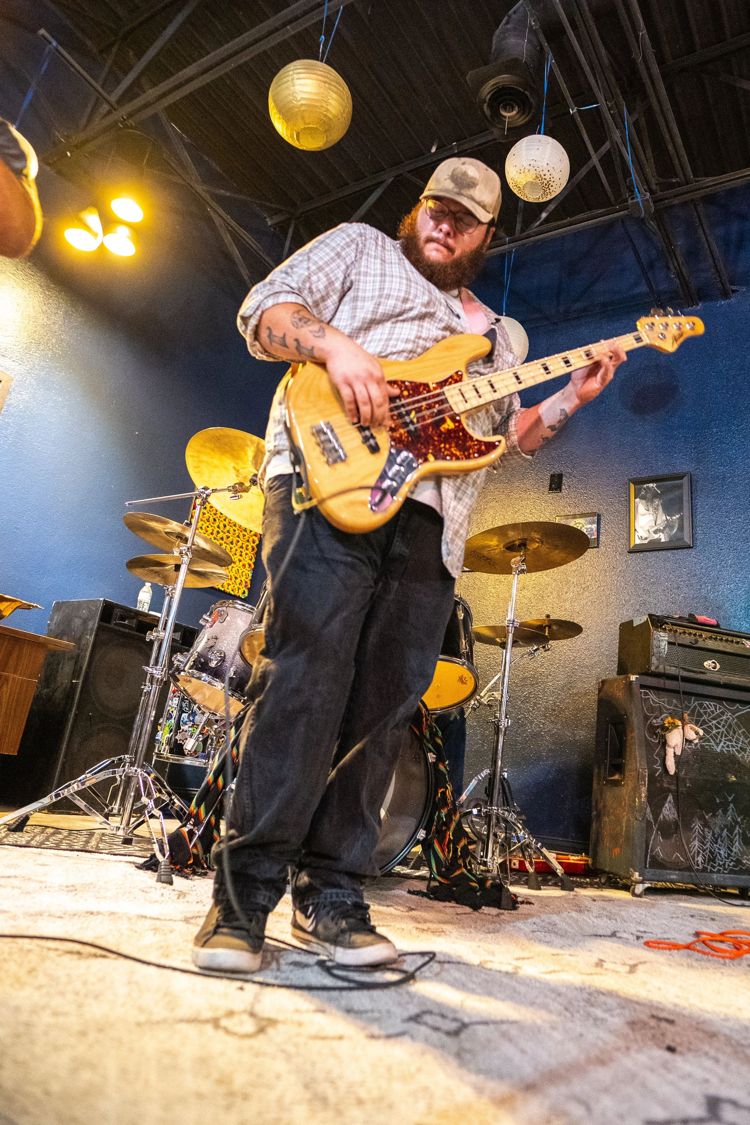 A man with tattoos, glasses, and a beard playing a yellow bass guitar on stage indoors, with drum set and speakers in the background, and decorative paper lanterns hanging from the ceiling.