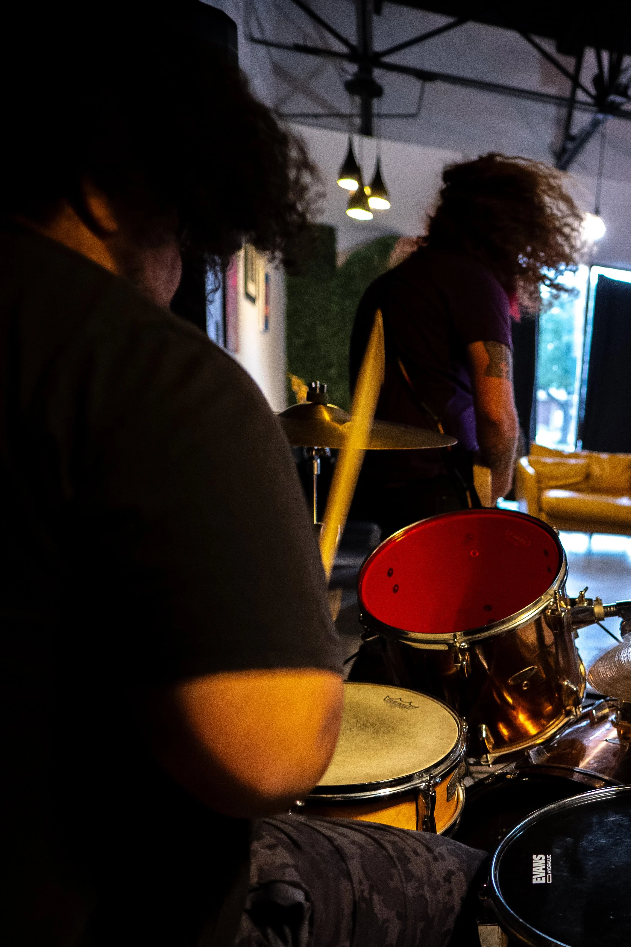 Person playing a drum set in a dimly lit room with a window in the background and modern hanging lights on the ceiling.