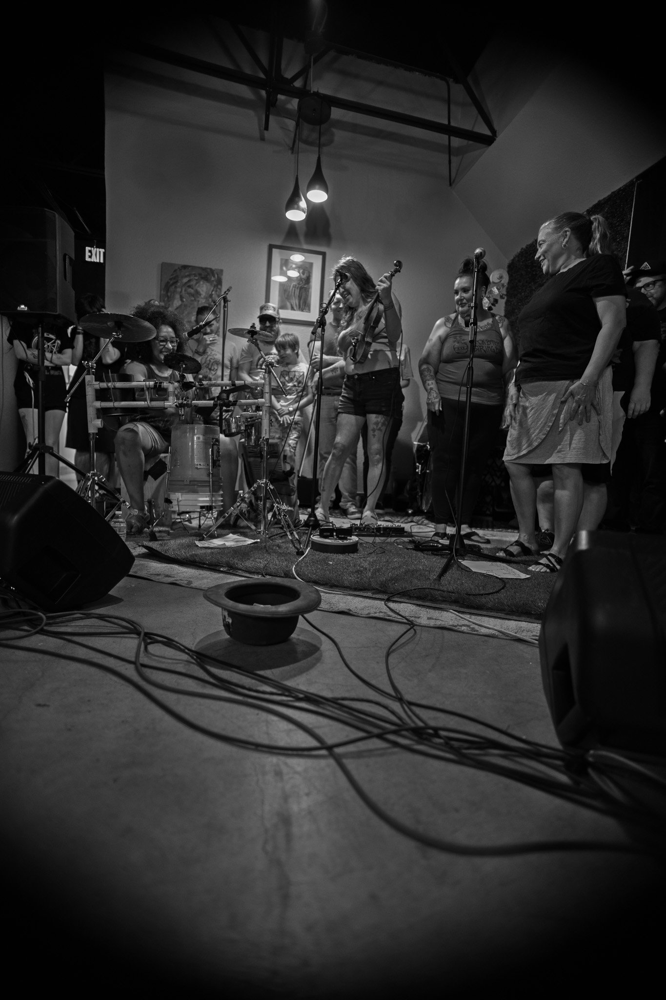 Black and white photo of a group of women performing on a stage, with musical instruments and microphones, in an intimate indoor venue with artwork on the wall.
