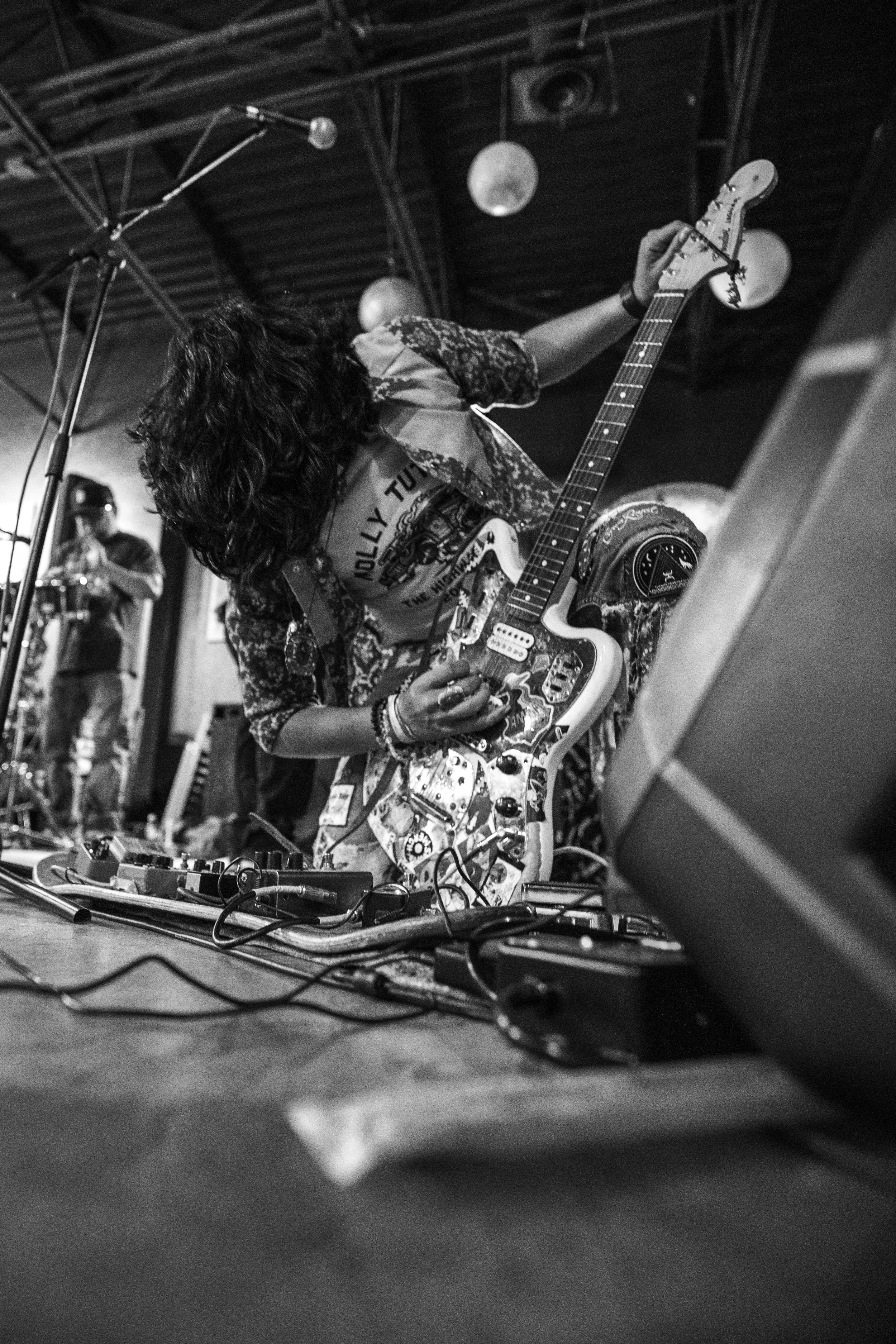 Musician with long curly hair playing electric guitar on stage, wearing a T-shirt and patterned jacket, with a man in the background and stage equipment visible.