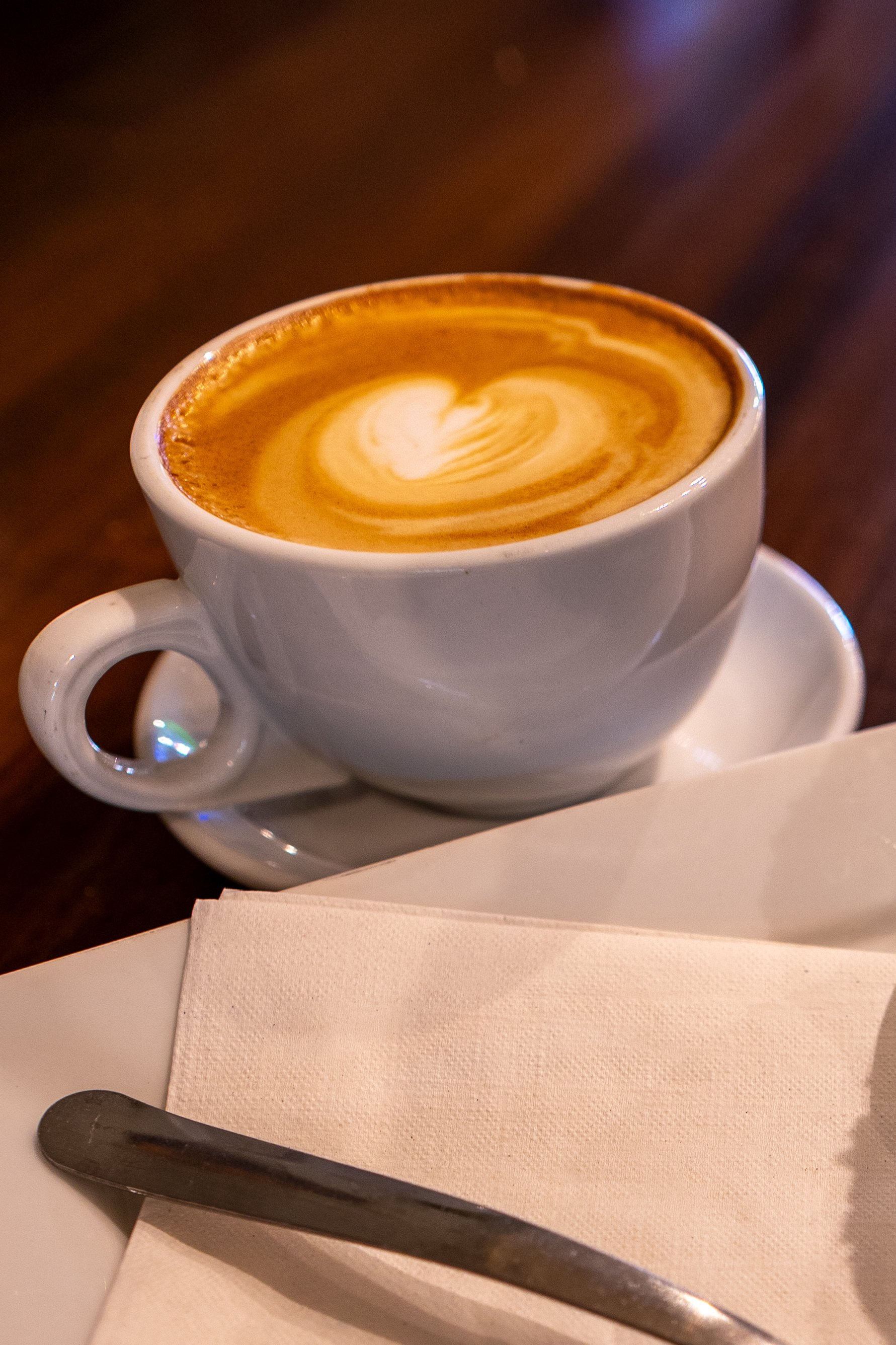 A cup of cappuccino with latte art, a white plate, a white napkin, and a spoon on a table.