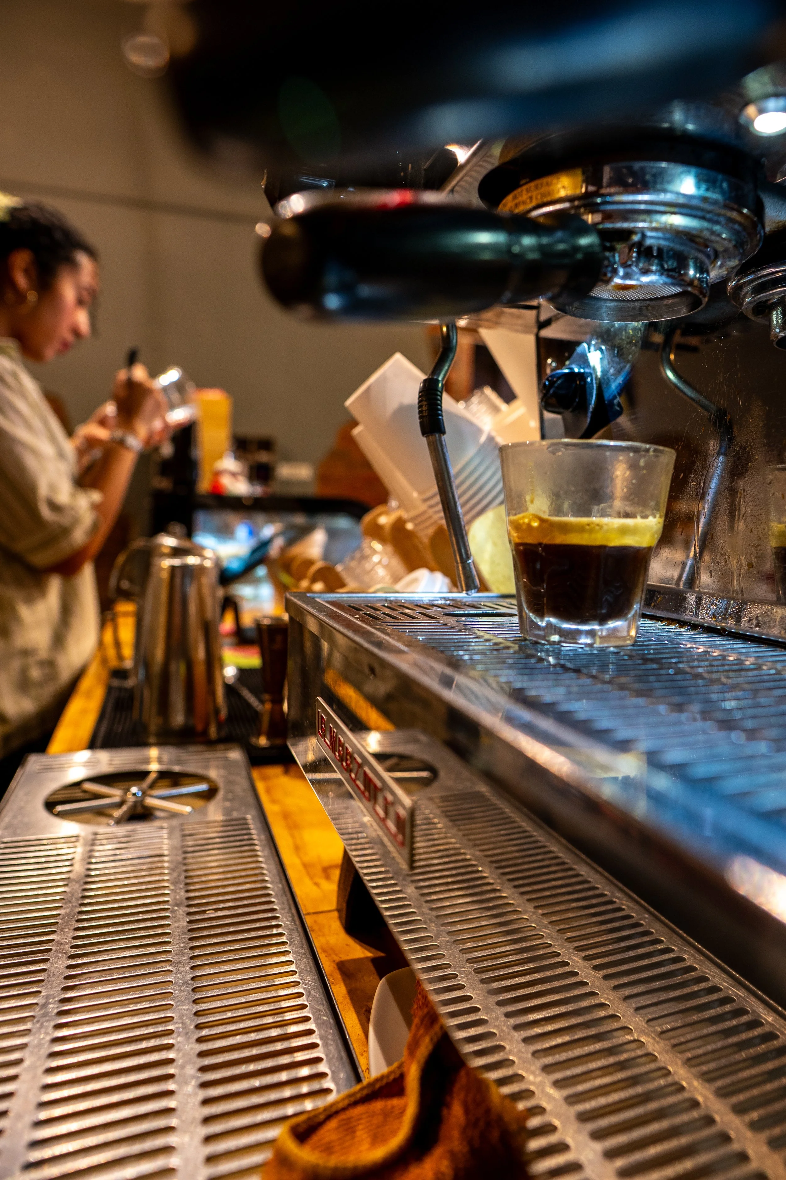 Close-up view of a coffee brewing machine with a glass of coffee placed underneath the dispenser, and a woman in the background preparing coffee in a coffee shop.