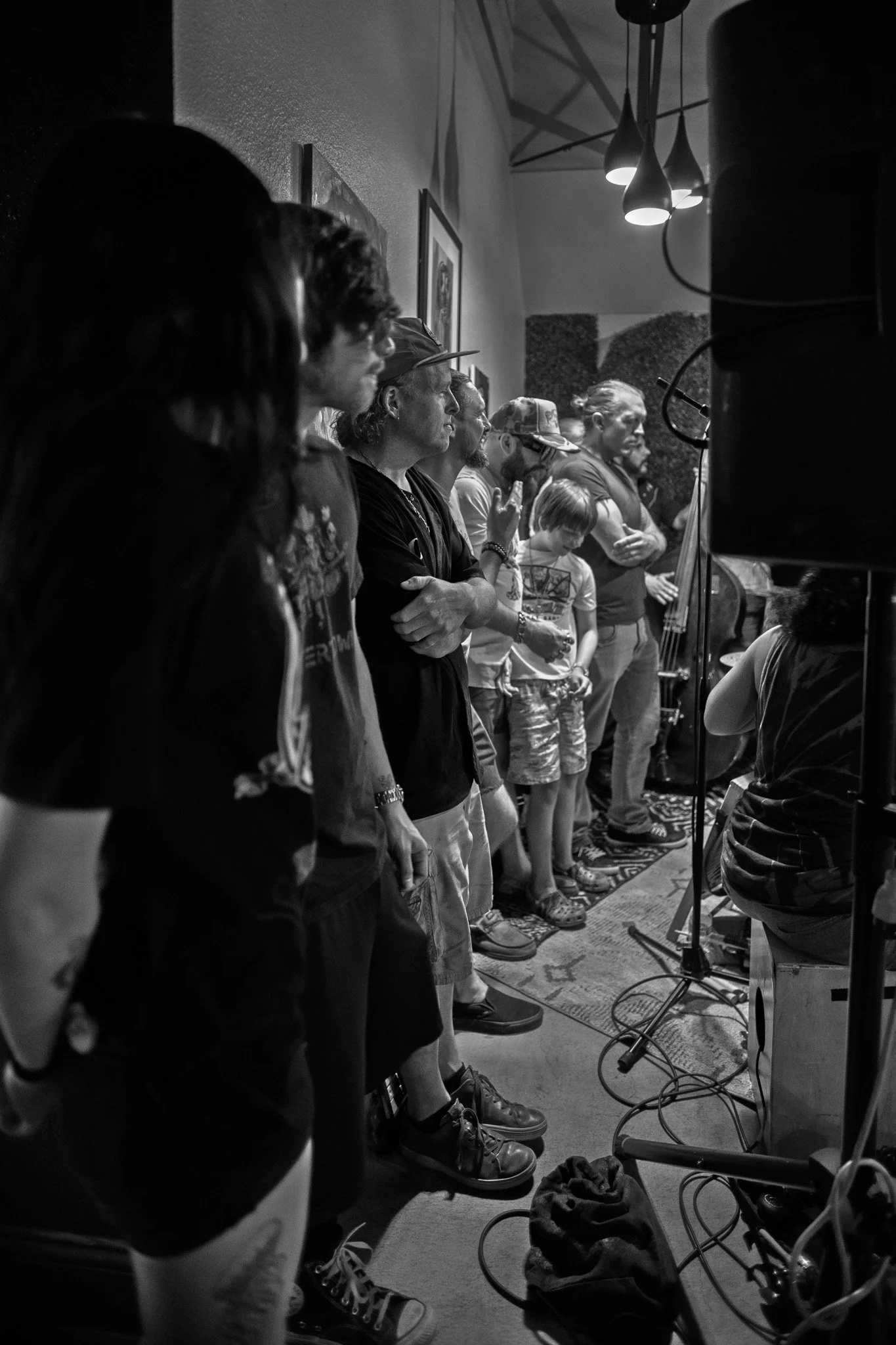 People standing in line inside a room, some with arms crossed, listening to a person speaking while holding a drumstick, with musical instruments in the background.