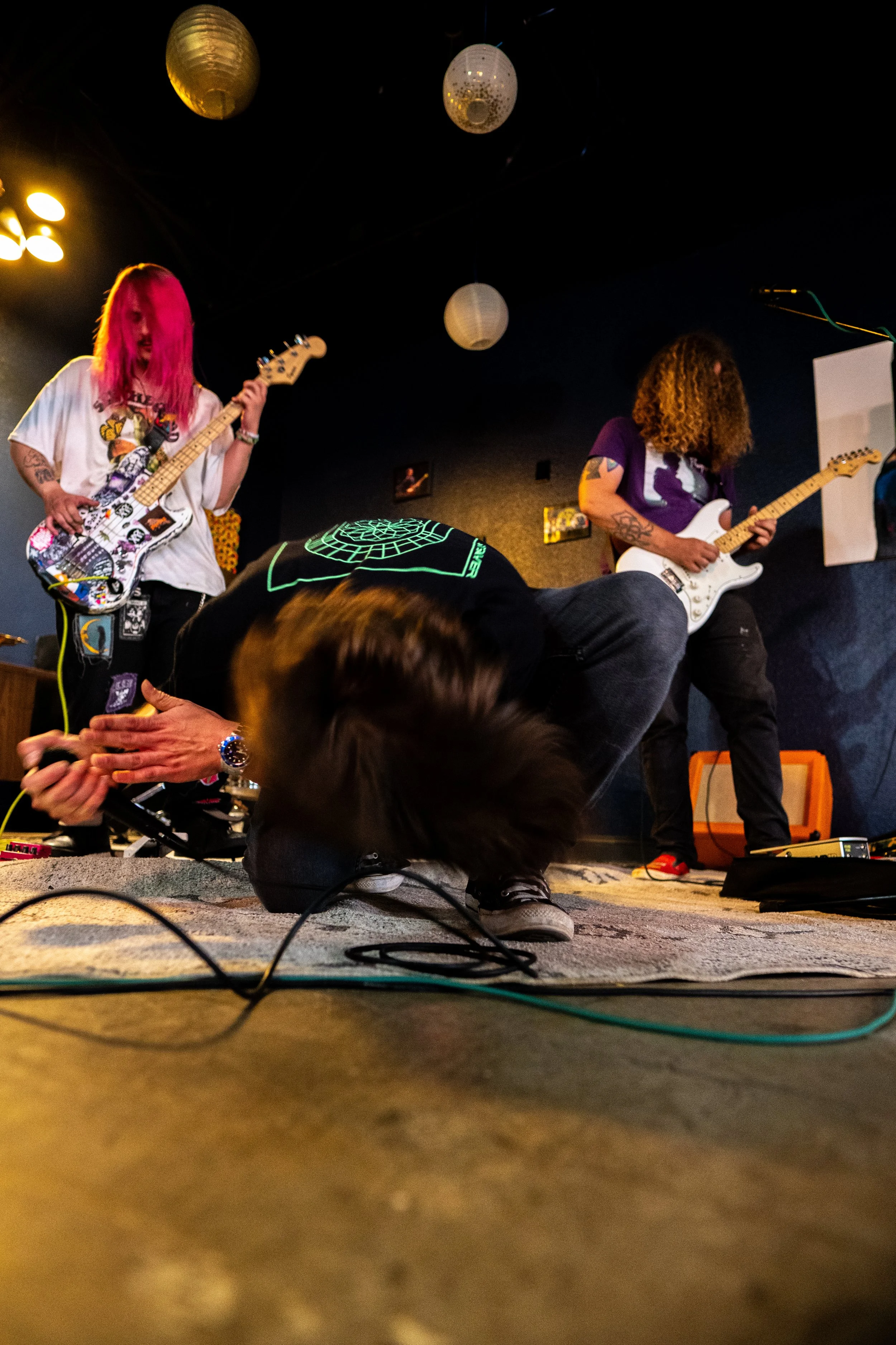 Two musicians performing on stage, one with pink hair playing bass guitar and the other with curly hair playing an electric guitar, with a person bending down in front, possibly adjusting equipment, in a dimly lit venue with hanging paper lanterns.