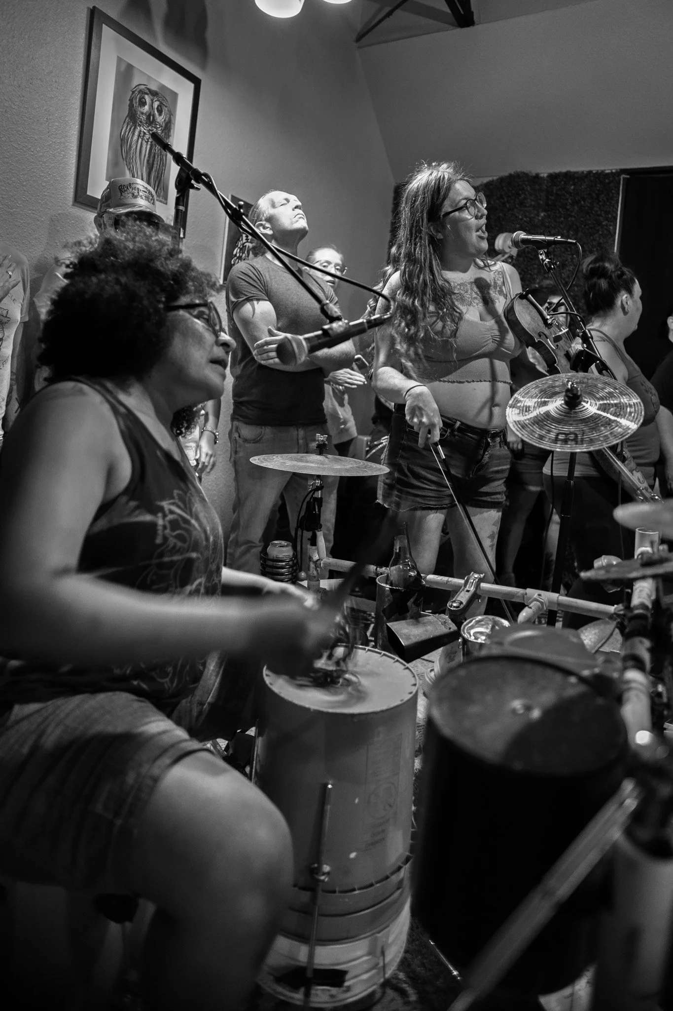 Black and white photo of a group of people at a music performance, including a woman with long wavy hair playing a violin, a woman with curly hair wearing glasses and a tank top, and a drummer in the foreground. The setting appears to be indoors with