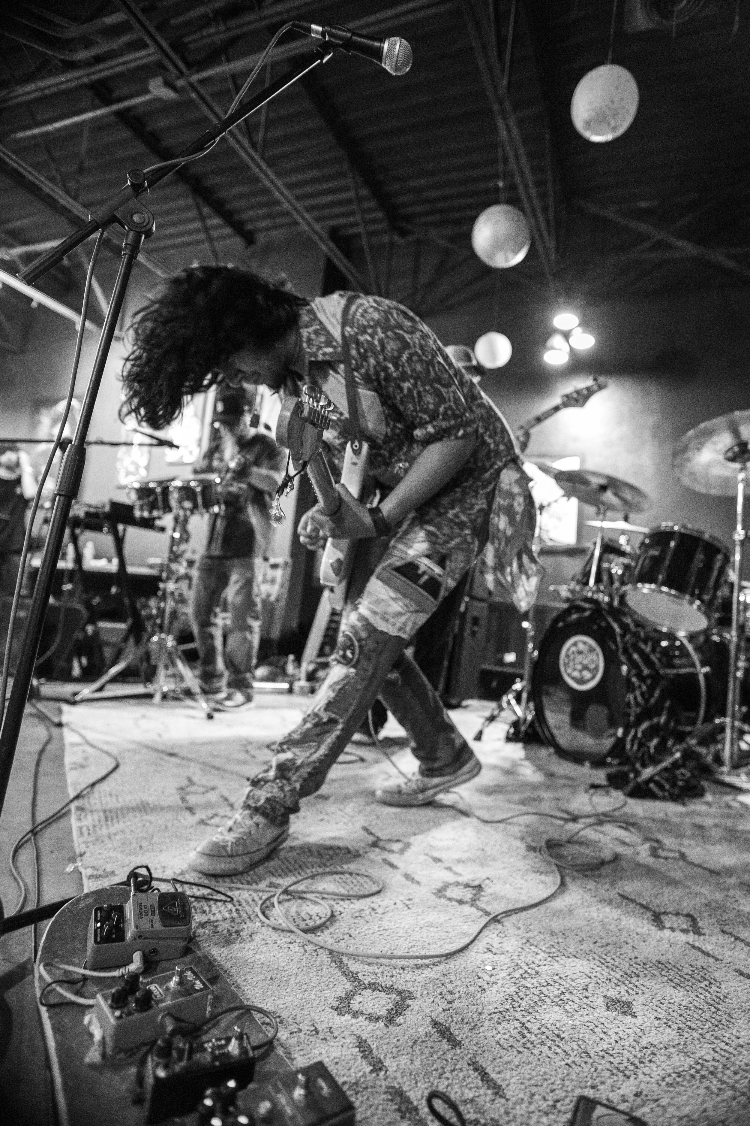 A black and white photo of a musician bending forward on stage, playing an electric guitar, with long hair covering their face. There are multiple drums behind them, microphone, and various guitar effects pedals on the floor.