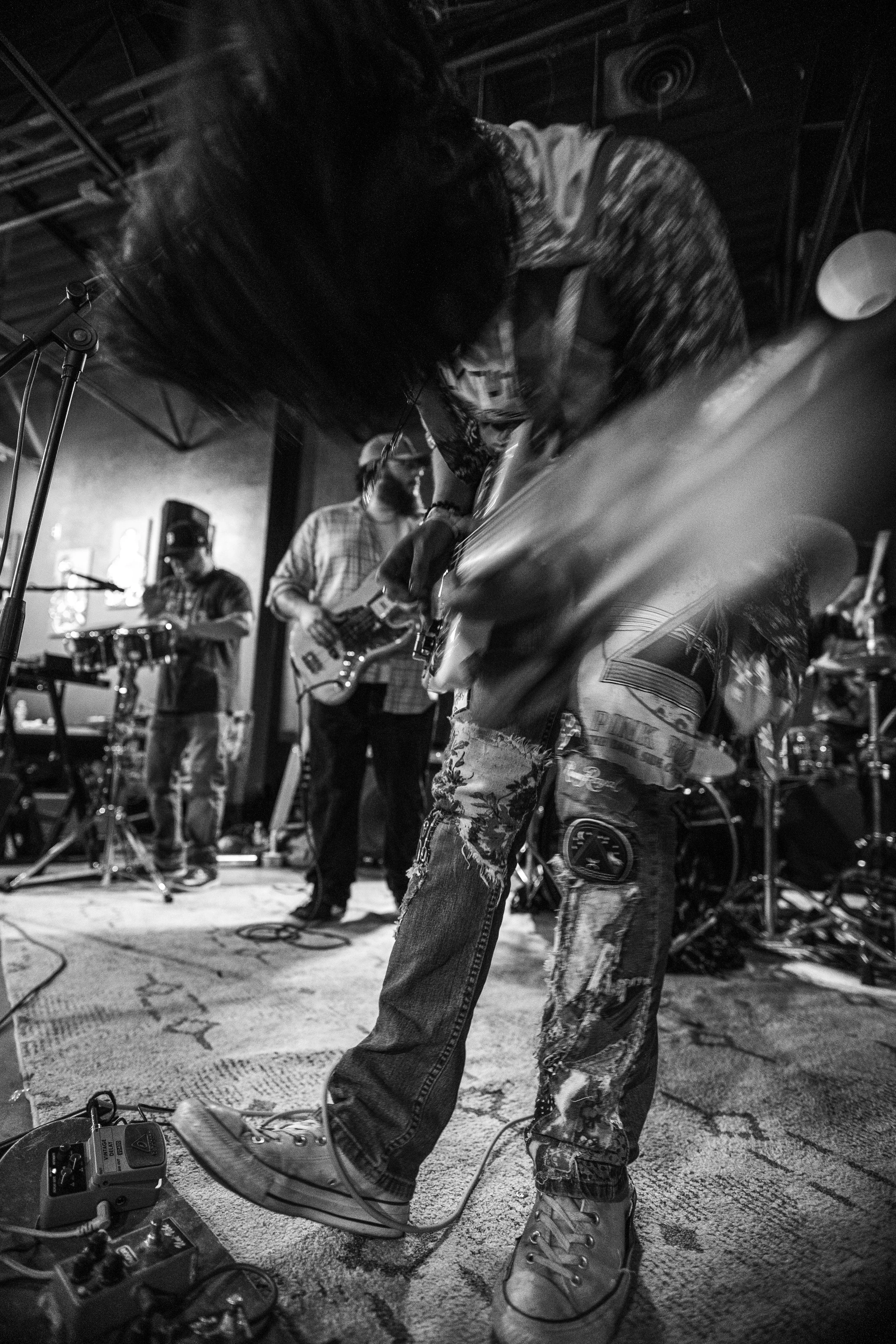 Musicians performing on stage, focusing on a guitarist's legs and shoes, surrounded by musical equipment and other band members in the background.