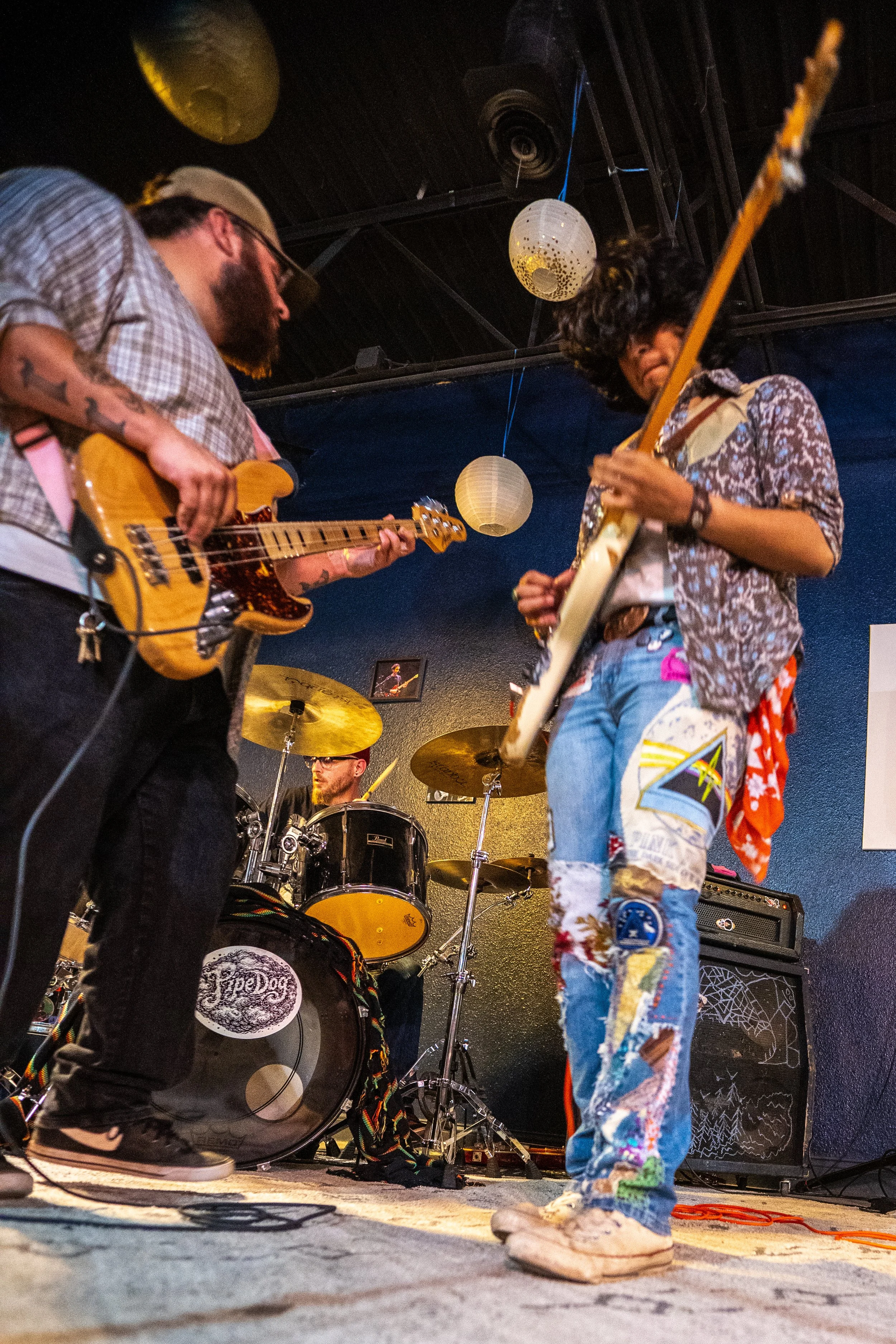 Two musicians, one with a guitar and the other with a bass guitar, are performing on a stage with a drummer in the background. The stage has hanging paper lanterns and wiring on the floor. The musician with the bass guitar is wearing colorful, patche