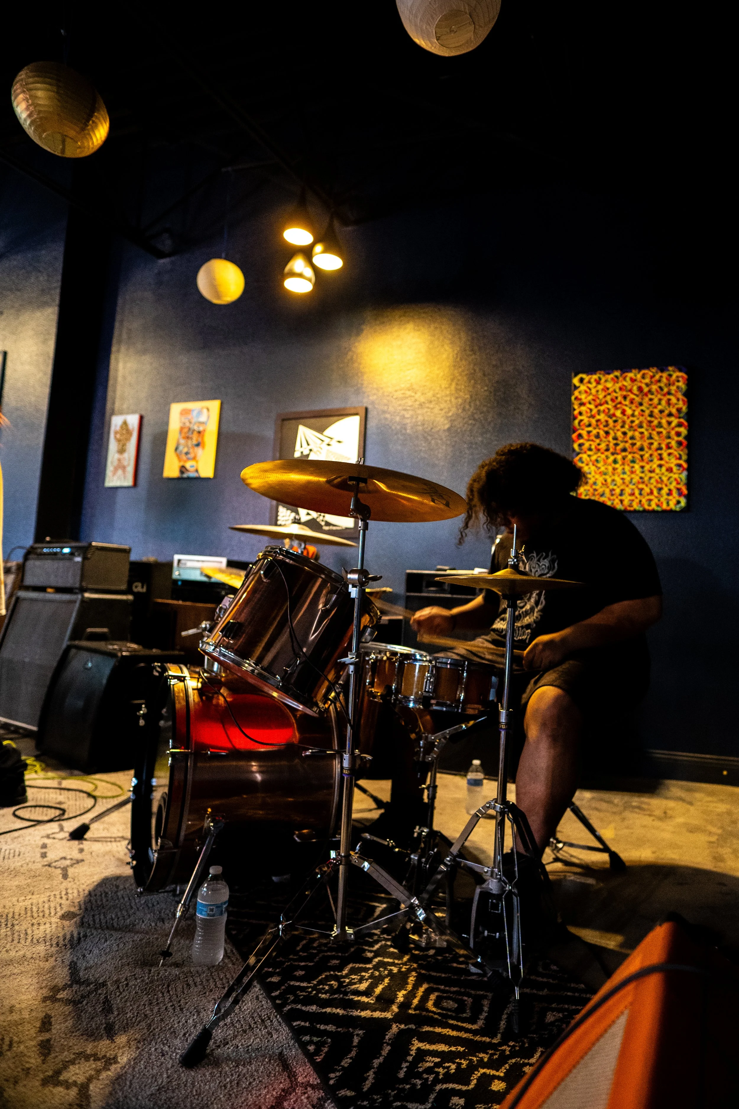 A person playing drums during a live music performance in a dark, decorated room with artwork on the wall and hanging paper lanterns.
