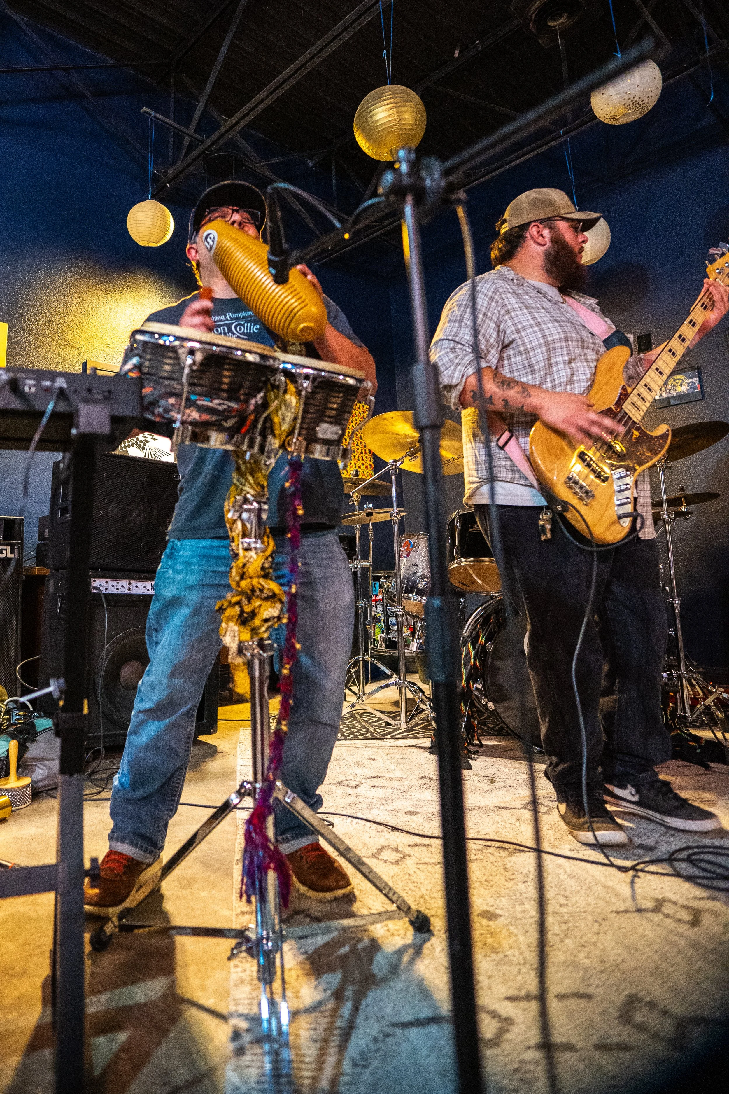 Two musicians performing on stage: one playing a combination of a musical instrument and a yellow lantern, and the other playing an electric bass guitar. The stage is decorated with hanging paper lanterns, and drum set is visible in the background.