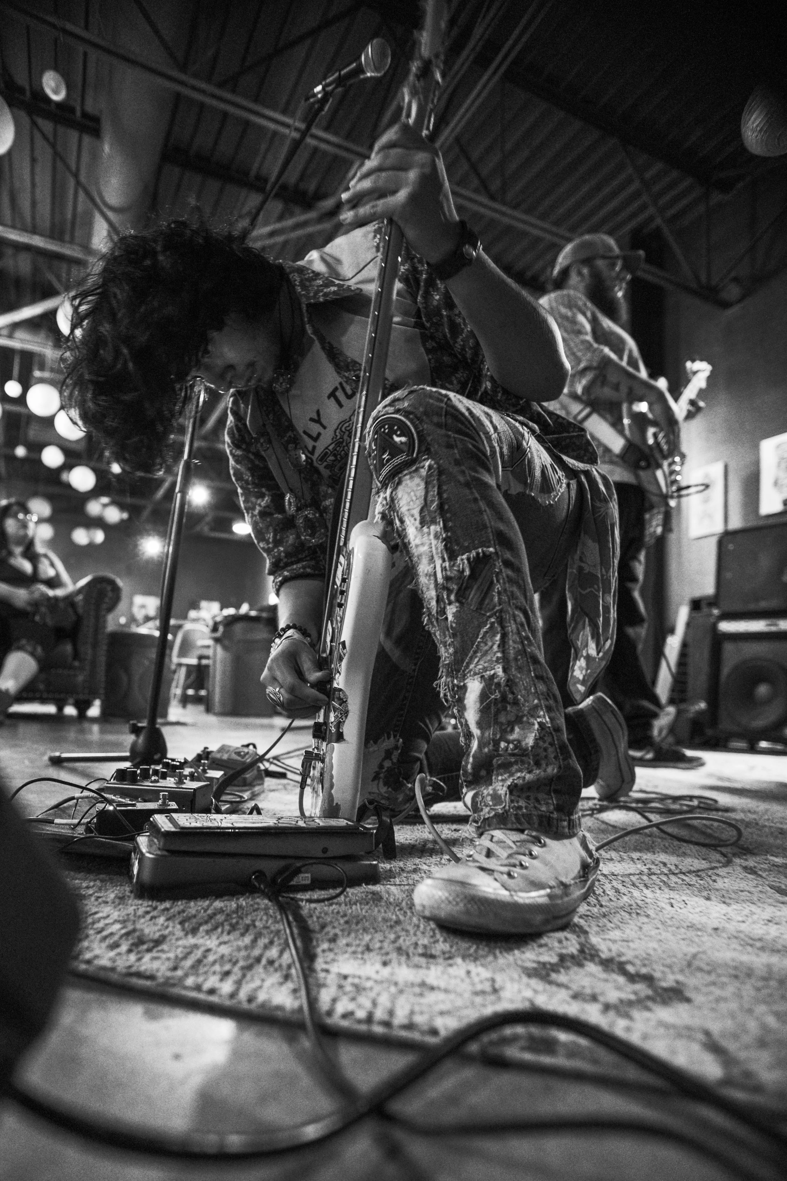 Two musicians sitting on the floor playing electric guitars on stage, surrounded by music equipment and cables in a performance setting.