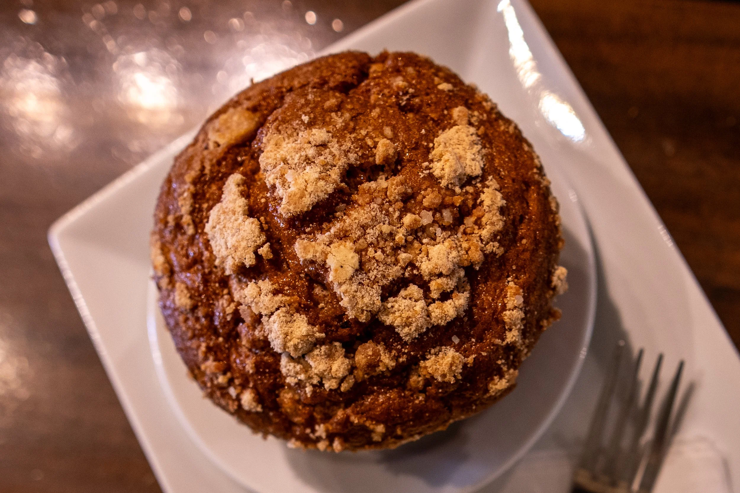 A close-up image of a muffin with a crumbly topping on a small white square plate.